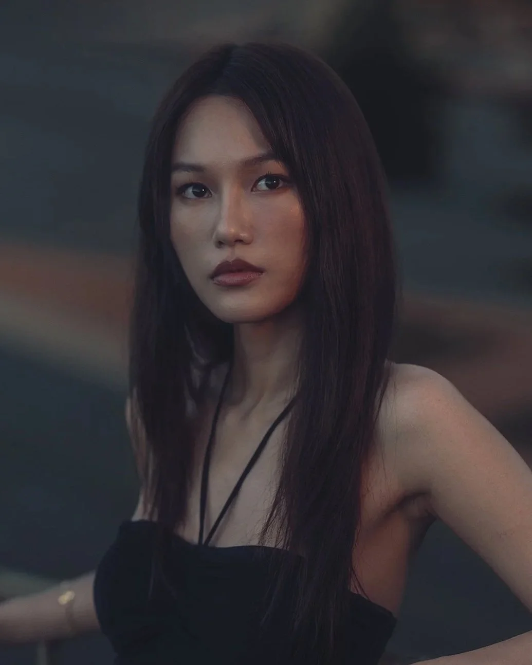 A woman with long dark hair and a serious expression wearing a black strapless top, posing outdoors during dusk or nighttime.