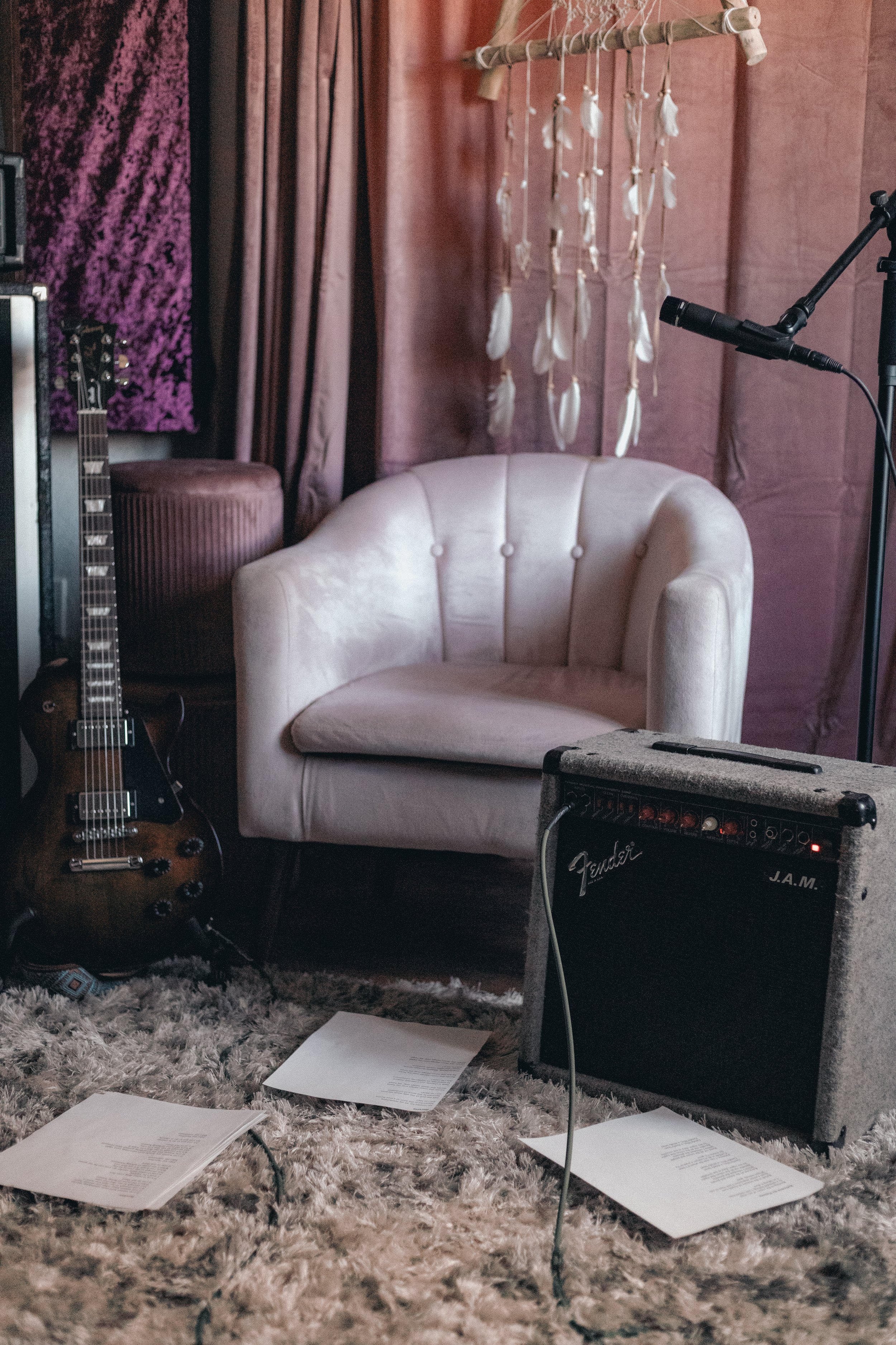 A cozy music studio corner featuring a white velvet armchair, an electric guitar on the floor, a Fender guitar amplifier with a cable, a few sheets of paper on a shaggy rug, a microphone on a stand, and a decorative feather hanging from a rod on a pink wall.