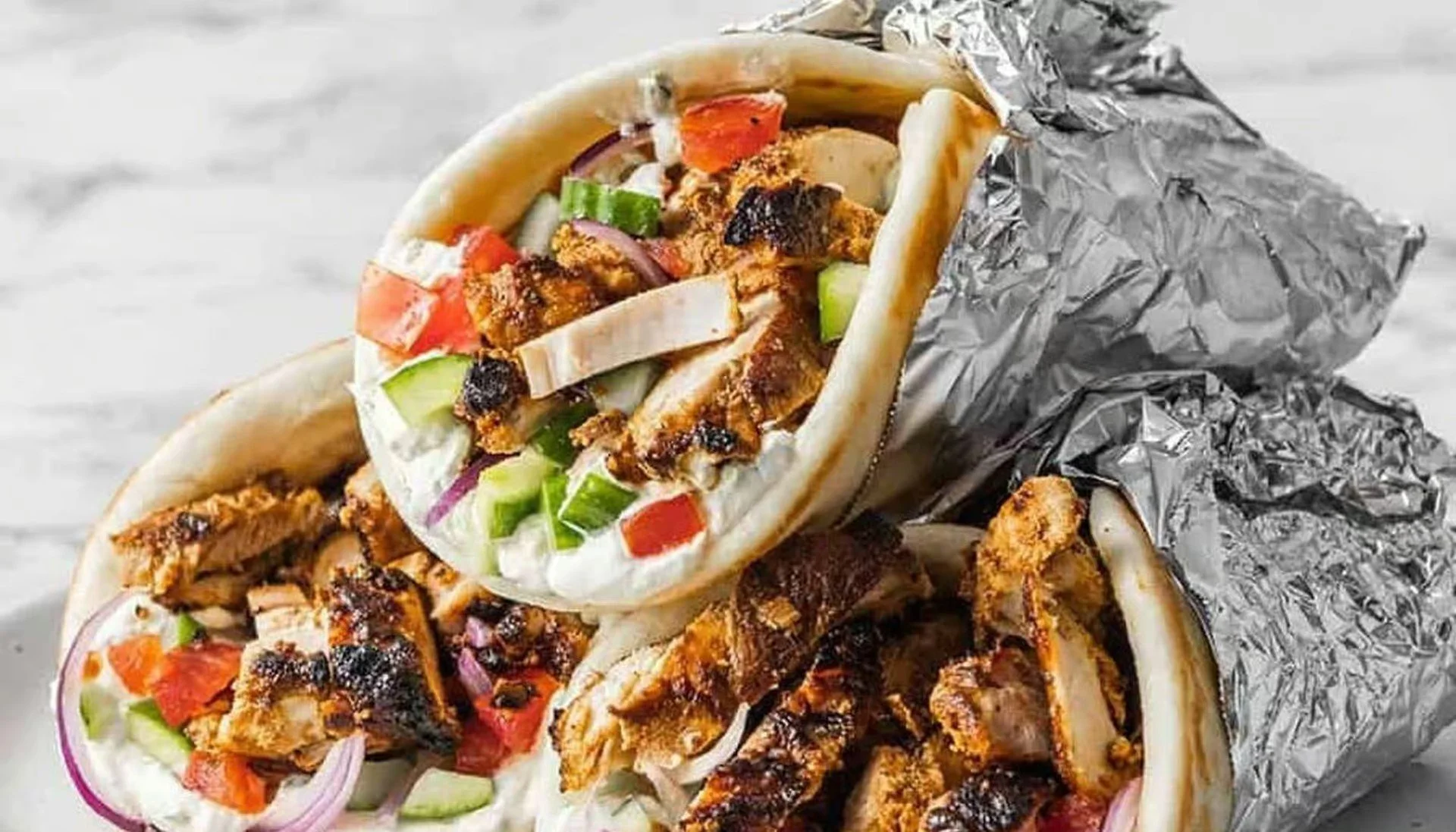Two chicken shawarma wraps partially unwrapped, showing grilled chicken, diced tomatoes, cucumbers, onions, and lettuce inside, with foil wrapping.