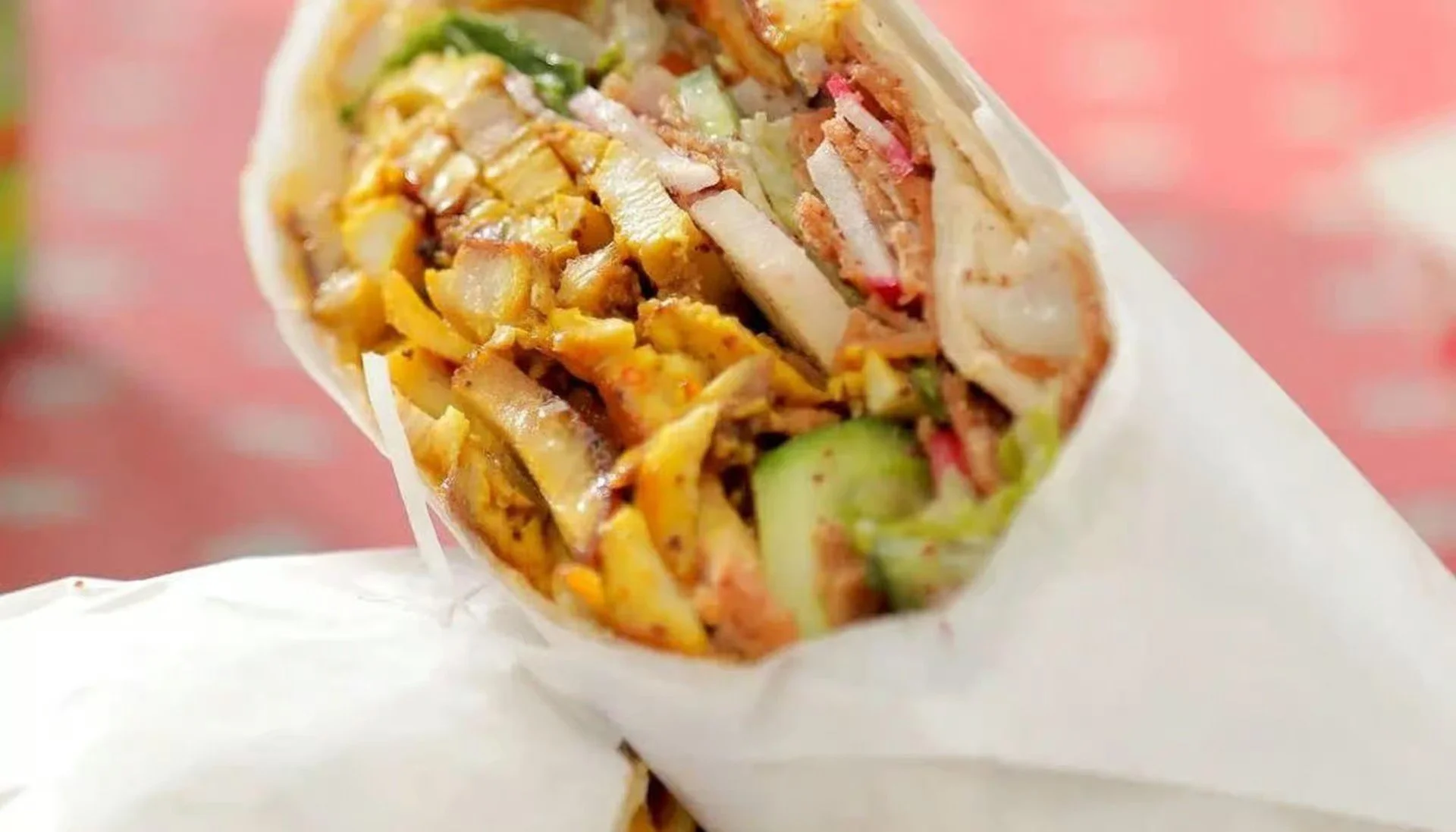 Close-up of a chopped Mexican burrito with visible ingredients including lettuce, meat, cheese, and sauce wrapped in a white tortilla, with a pink background.