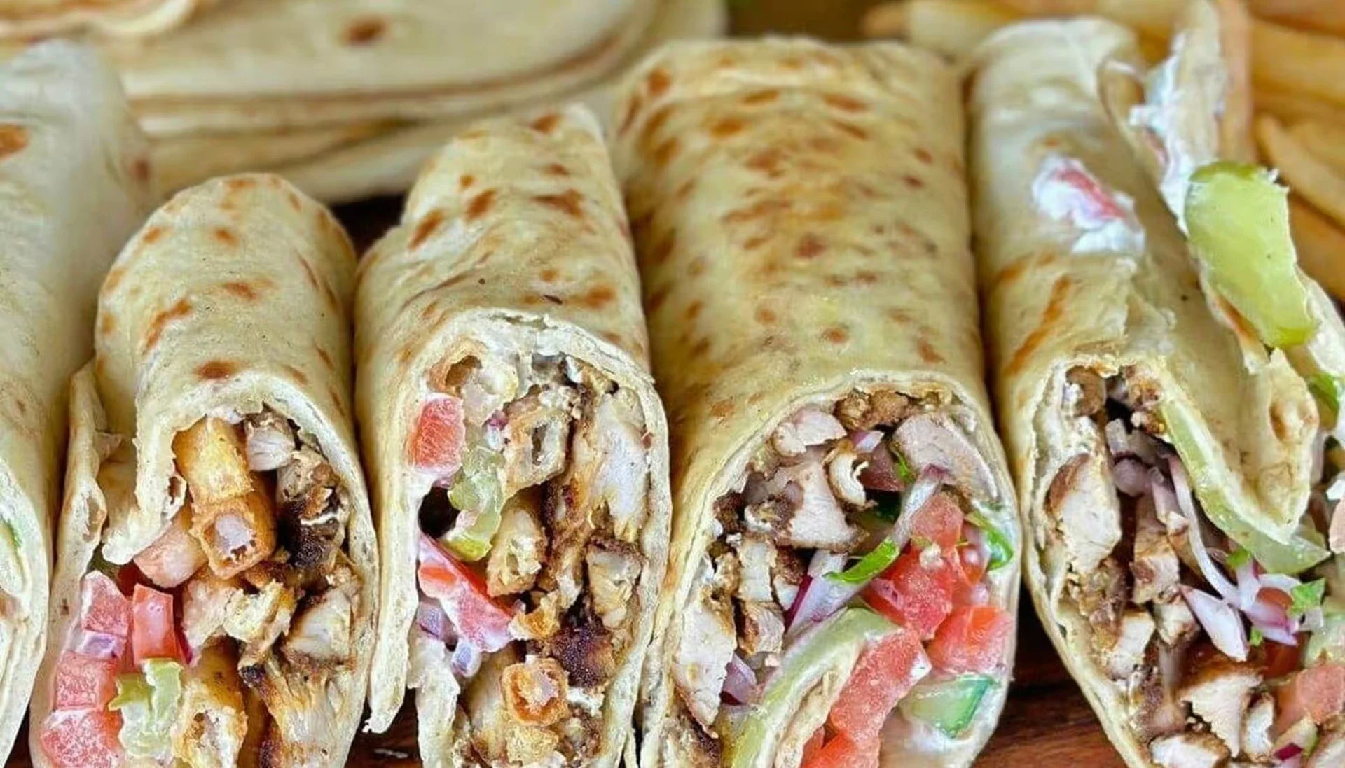 Close-up of four burritos with meat, lettuce, tomato, and cheese, wrapped in flour tortillas.