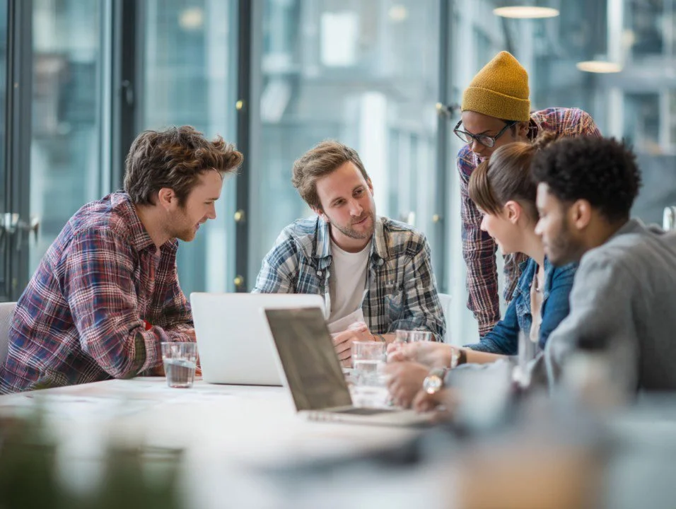 Group of six young adults working together at a conference room table with laptops and glasses of water.