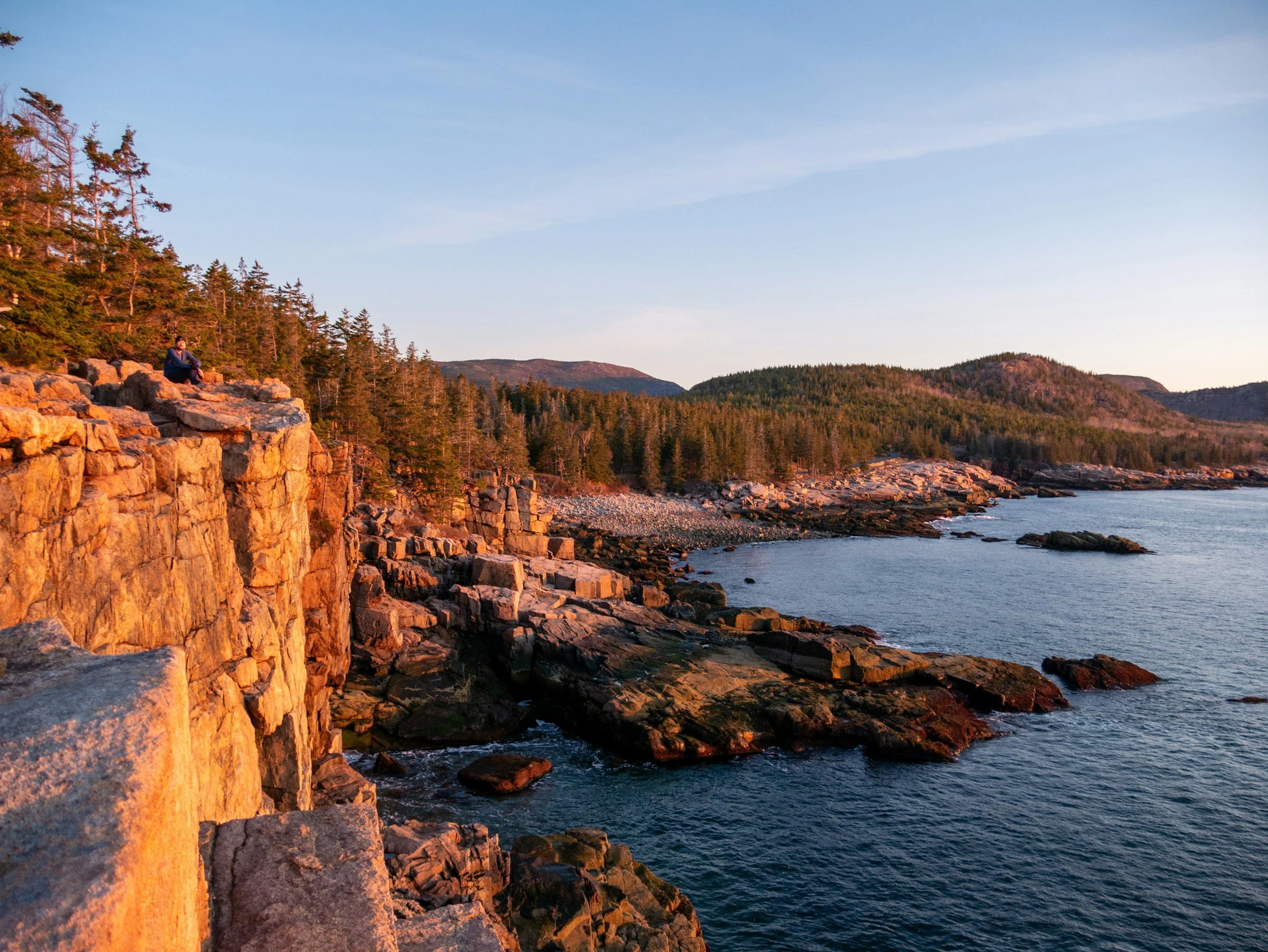 A person sitting on a rocky cliff overlooking the ocean during sunset, with forested hills in the background.