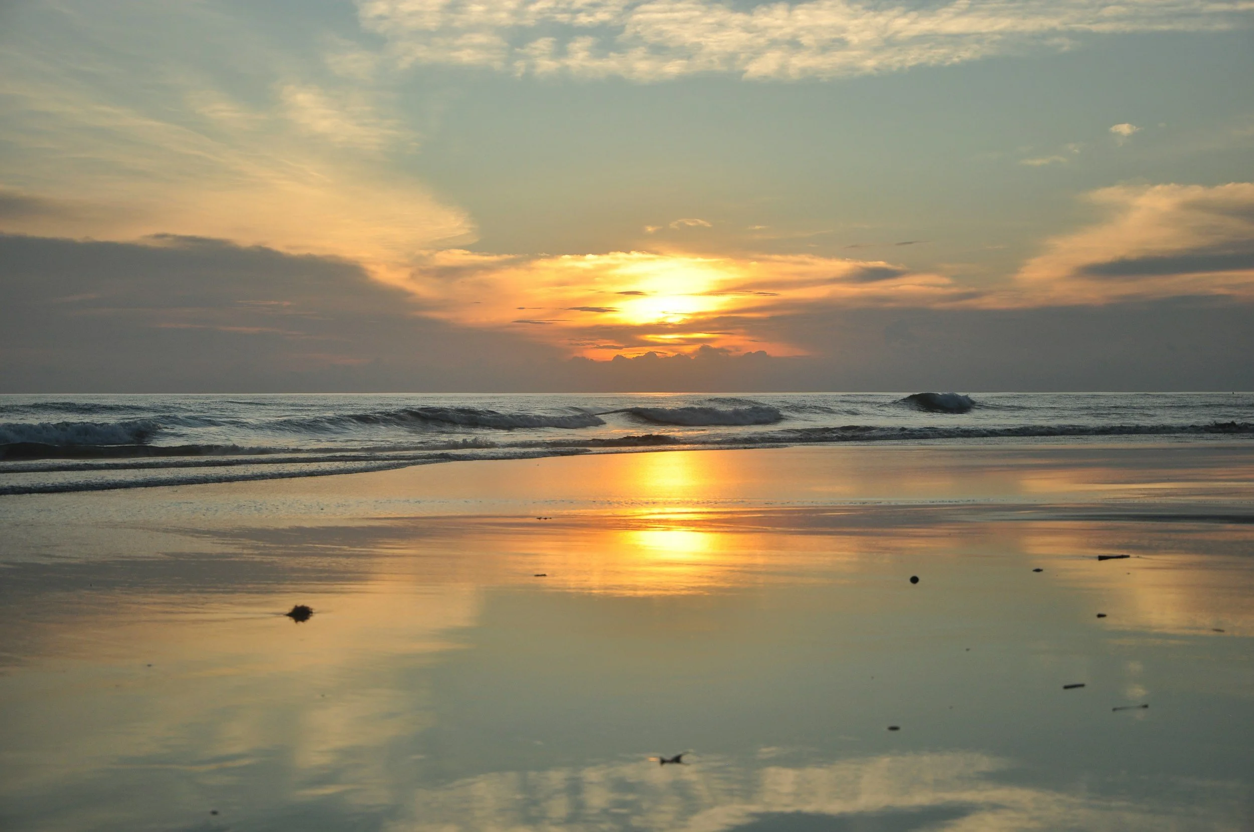 Sunset over the ocean with waves crashing onto the beach, reflected on wet sand, and a partly cloudy sky.