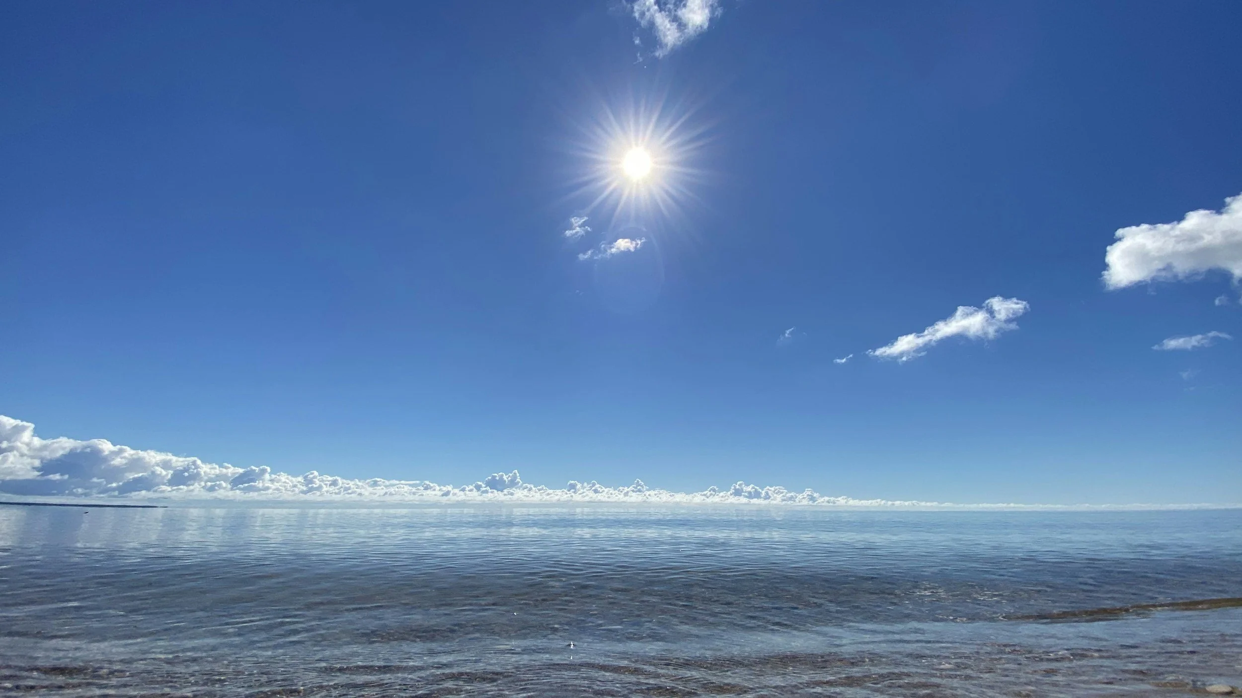 Clear blue sky with the sun shining brightly, a few clouds, calm ocean water, and a sandy shoreline in the foreground.