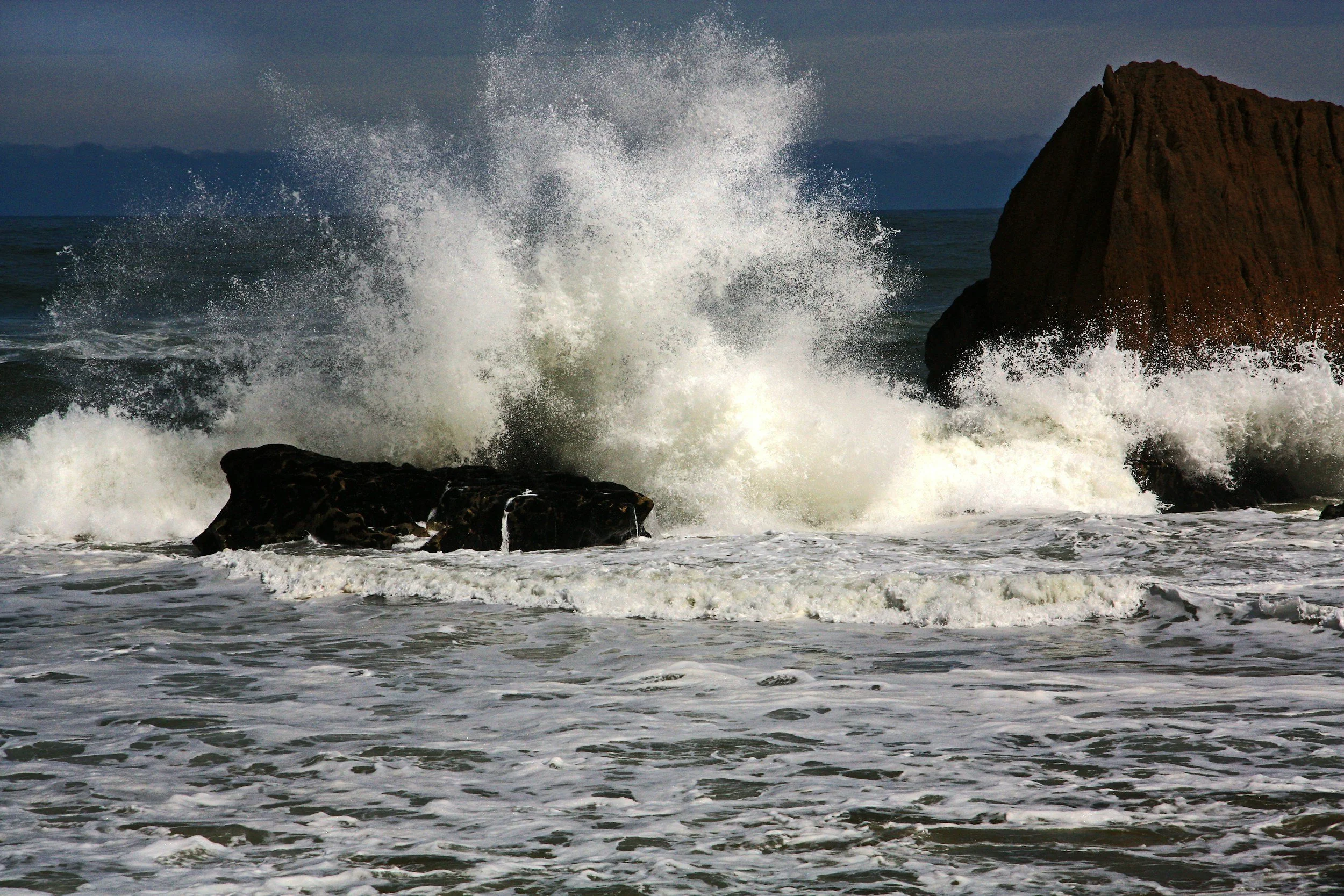 Waves crashing against rocks on the shoreline with a large rock formation in the background.