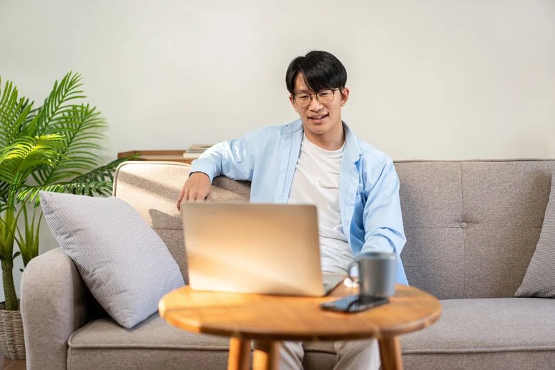 Young man with glasses in a light blue shirt on his laptop speaking with an online therapist in Florida on a cozy sofa with cushions, a coffee mug, and a plant in the background.