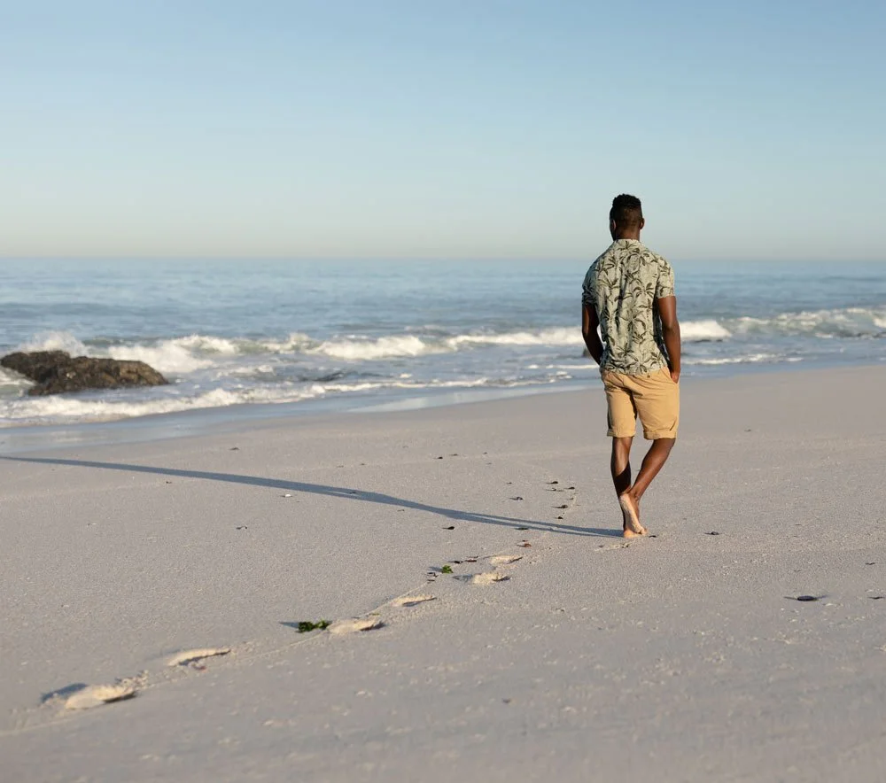 A man walking along the beach with his hands in his pockets, leaving footprints in the sand with the ocean and a clear sky in the background.