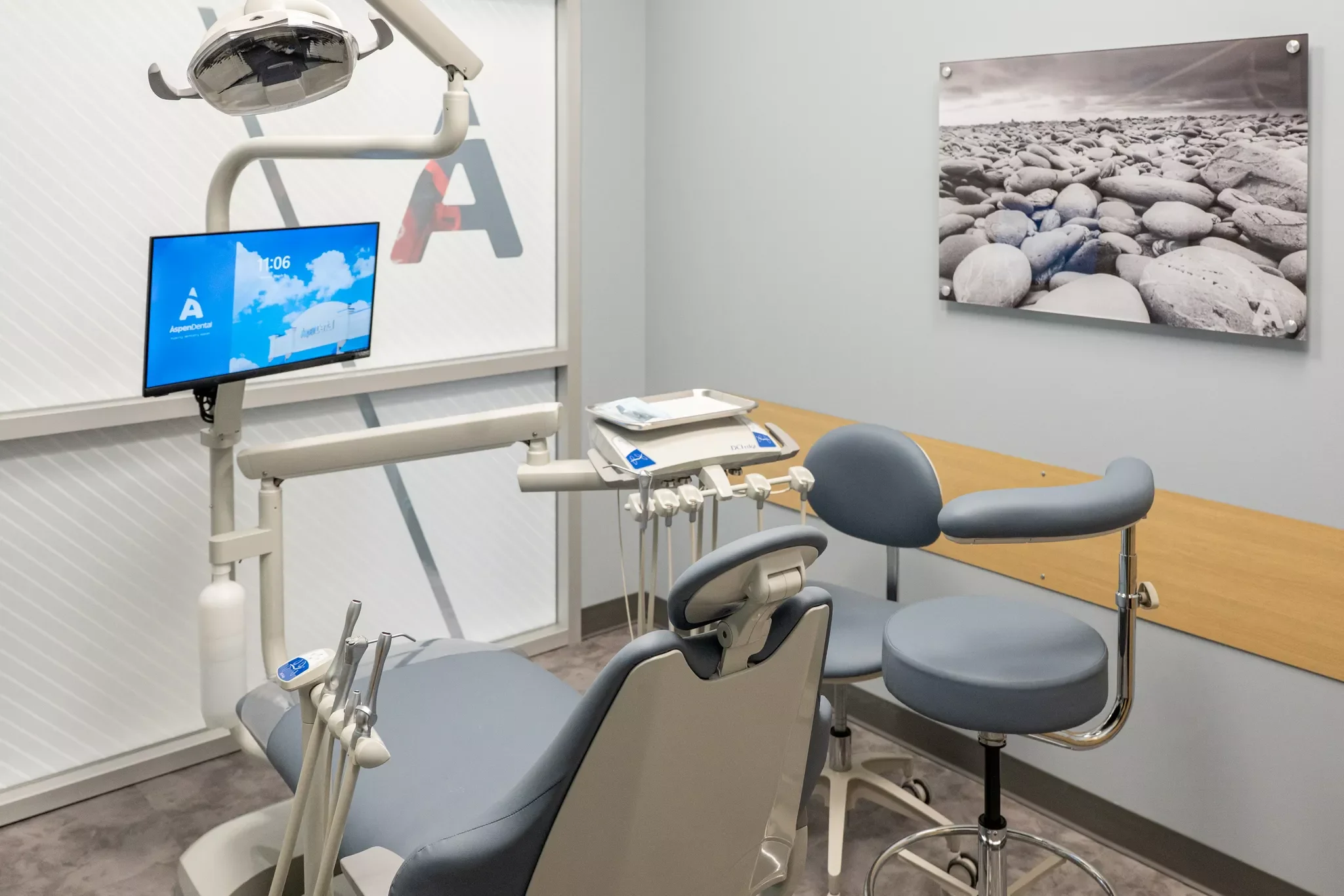 Dental examination room with dental chair, overhead light, monitor, and dental tools, with a stone beach picture on the wall.