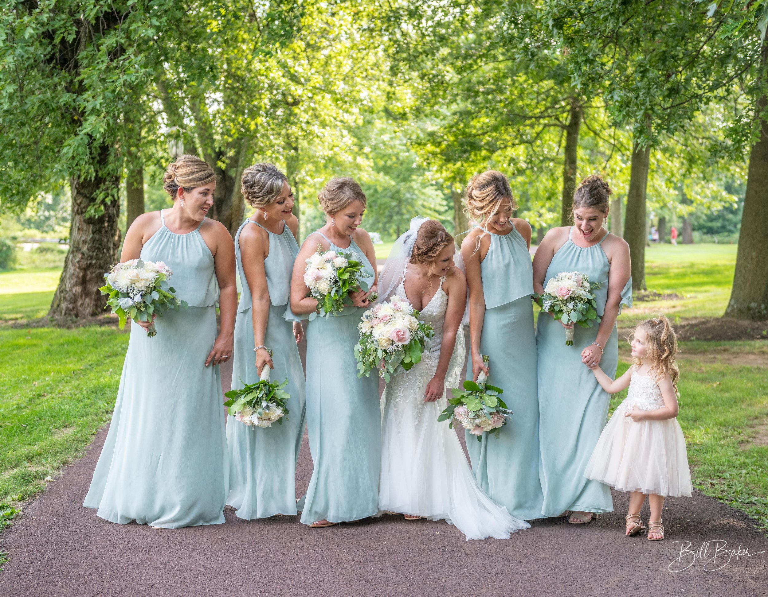 A bride and six bridesmaids, all holding bouquets, walking outdoors on a paved path in a park with green trees and grass, happily interacting.