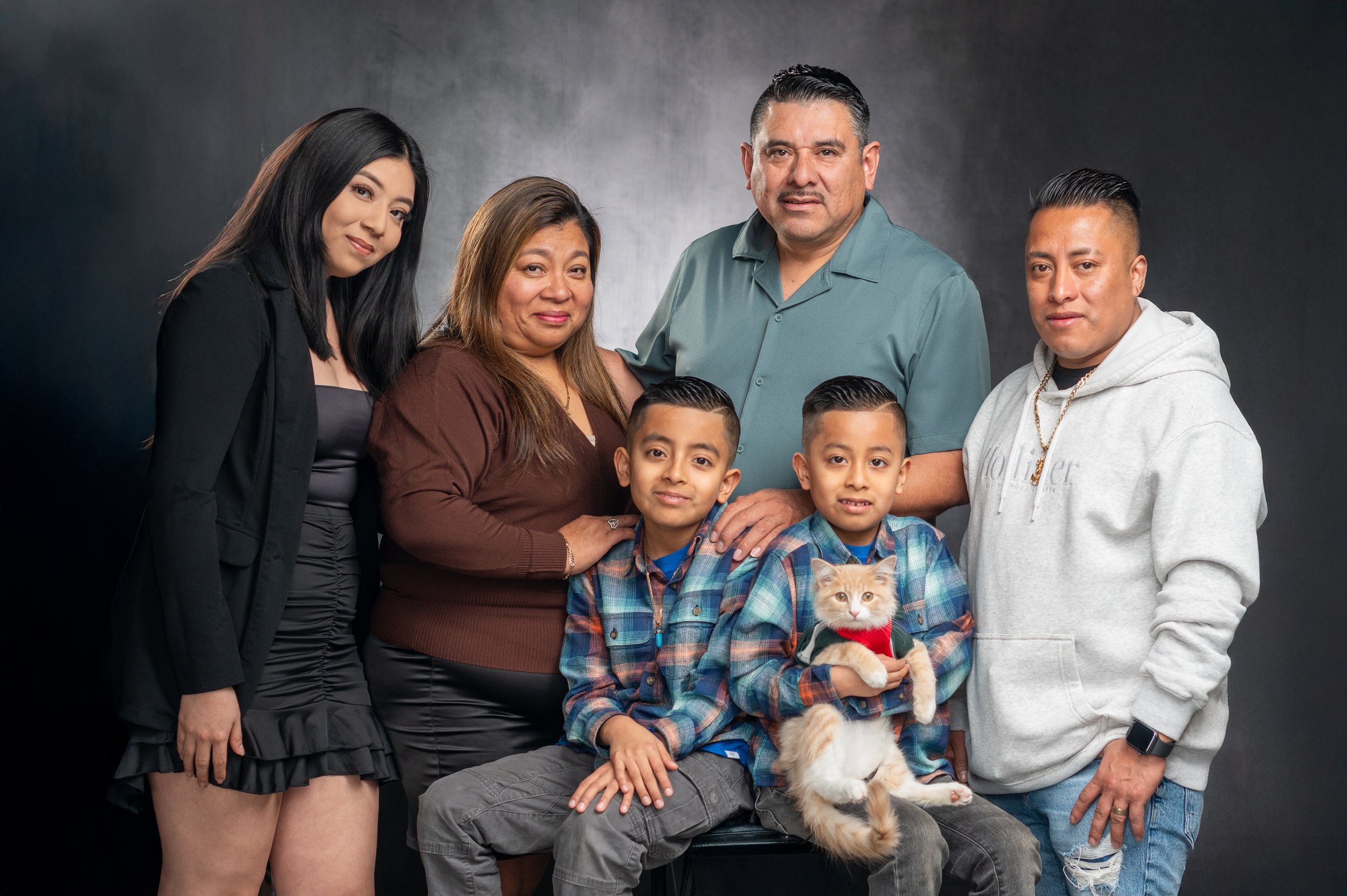 A multigenerational family portrait of seven members, including two young boys, two adult men, two adult women, and a cat, posing in front of a dark background.