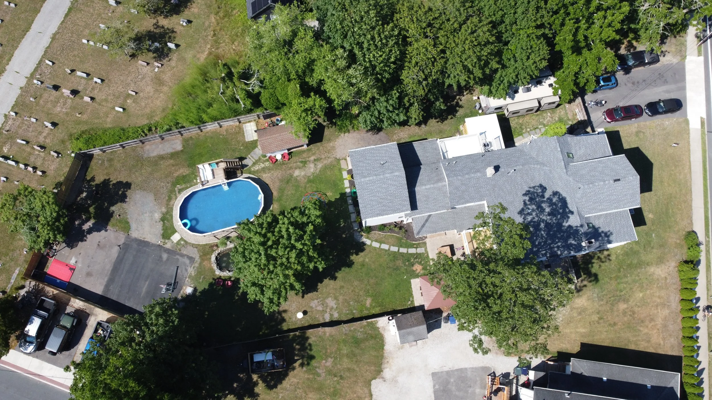 From an aerial view, the image shows a backyard with a swimming pool, trees, a house, a driveway with parked cars, a small shed, and a fenced area with headstones.