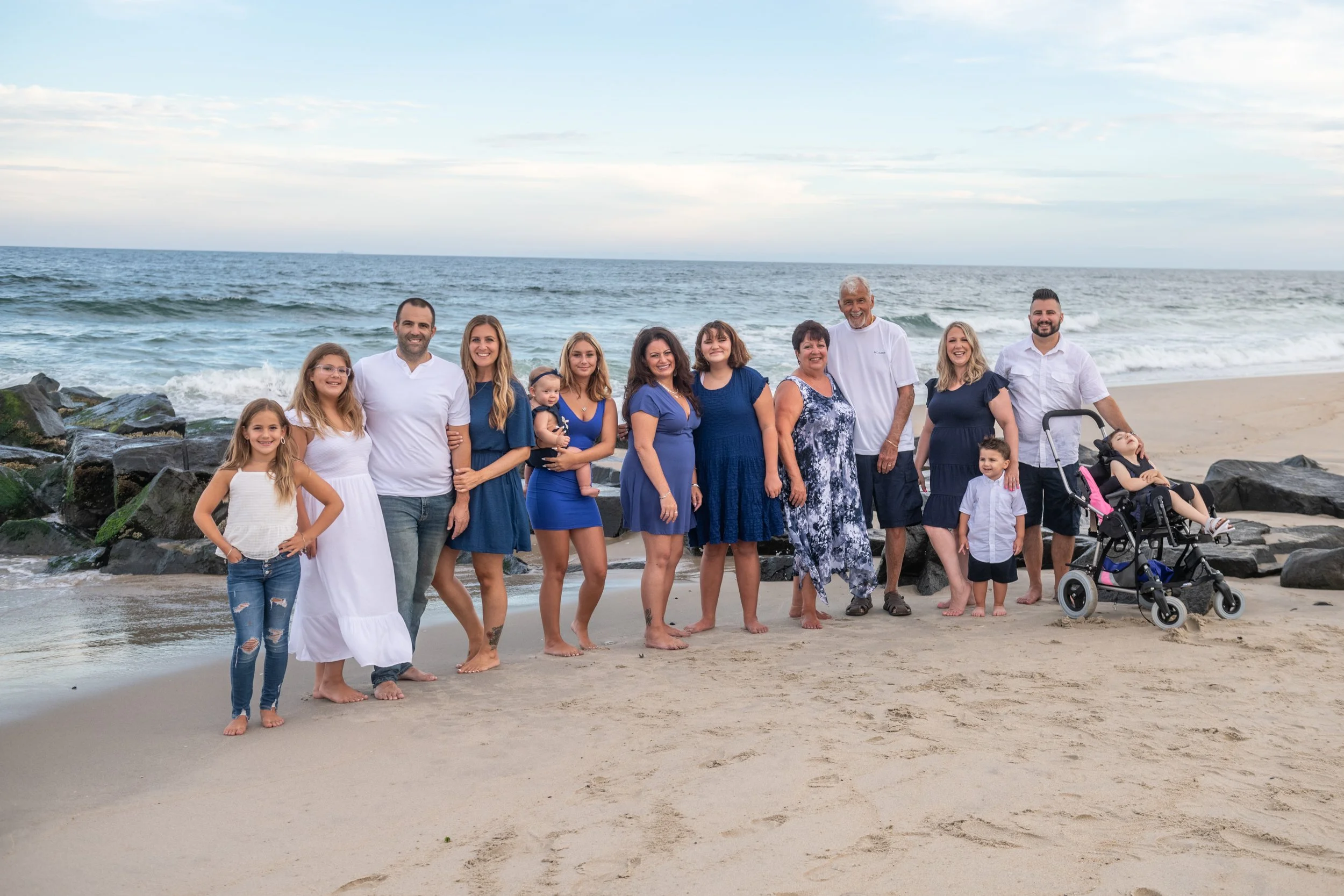 Family group photo on the beach featuring adults and children, including a woman in a white dress, a man in a white shirt, several women and girls in blue dresses, a woman in a patterned dress, and a man with a stroller, all standing in front of the ocean with rocks and sand.