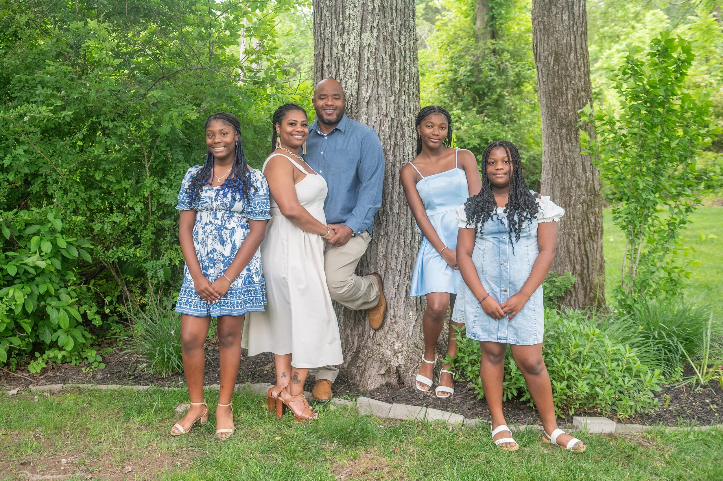 Family of six standing outdoors beside a large tree in a lush green garden, smiling and dressed in summer clothing.