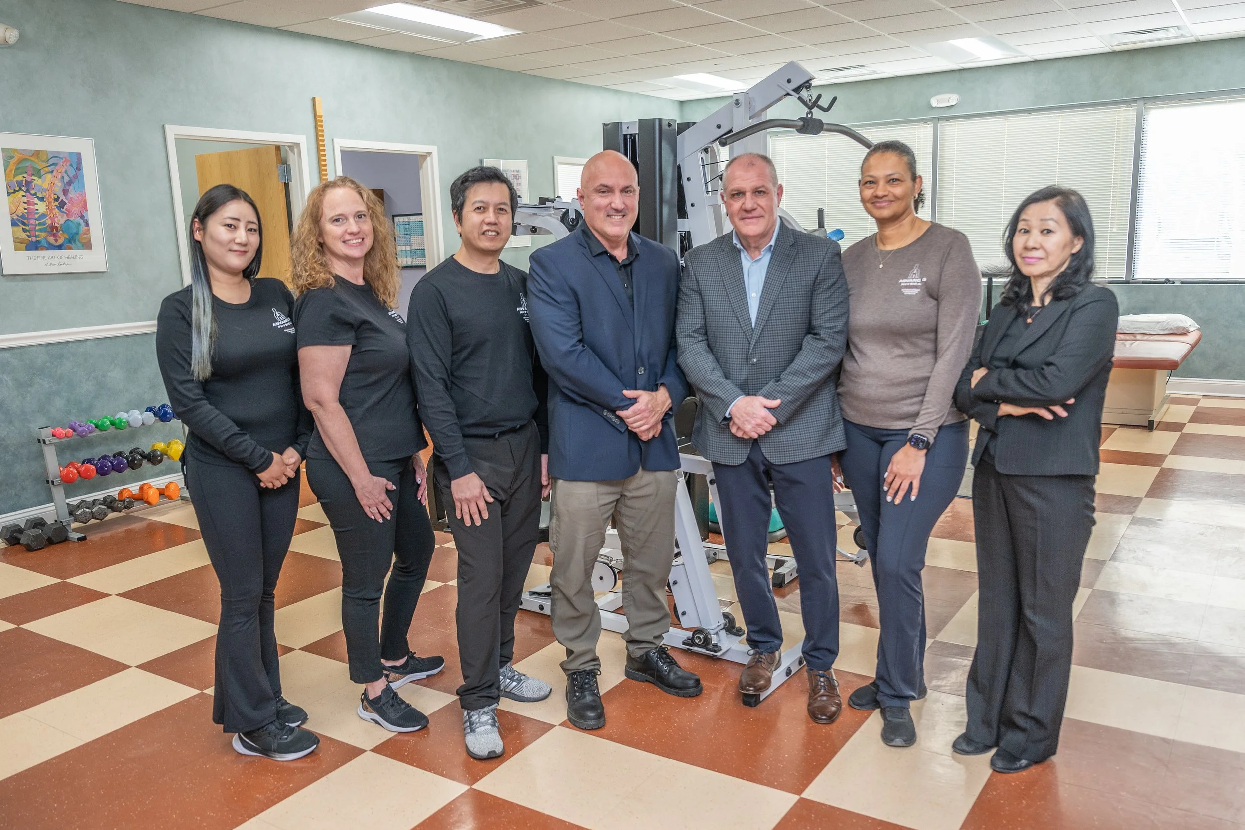 A group of seven people standing inside a physical therapy or rehabilitation clinic, with exercise equipment and a massage table visible in the background.