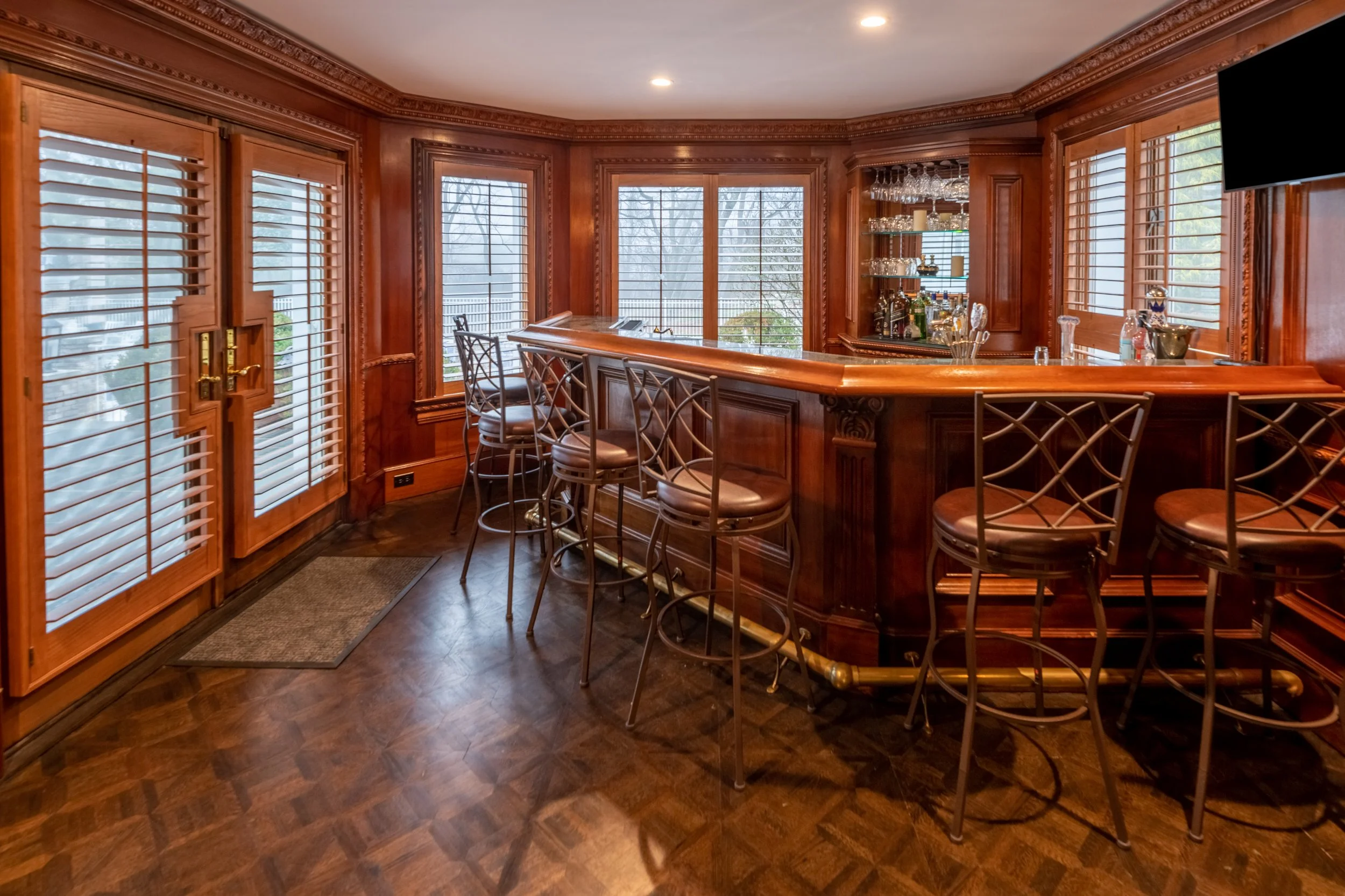 Wood-paneled home bar with a curved counter, topped with bottles and glasses, and several bar stools around it. Windows with wooden shutters show an outdoor scene.
