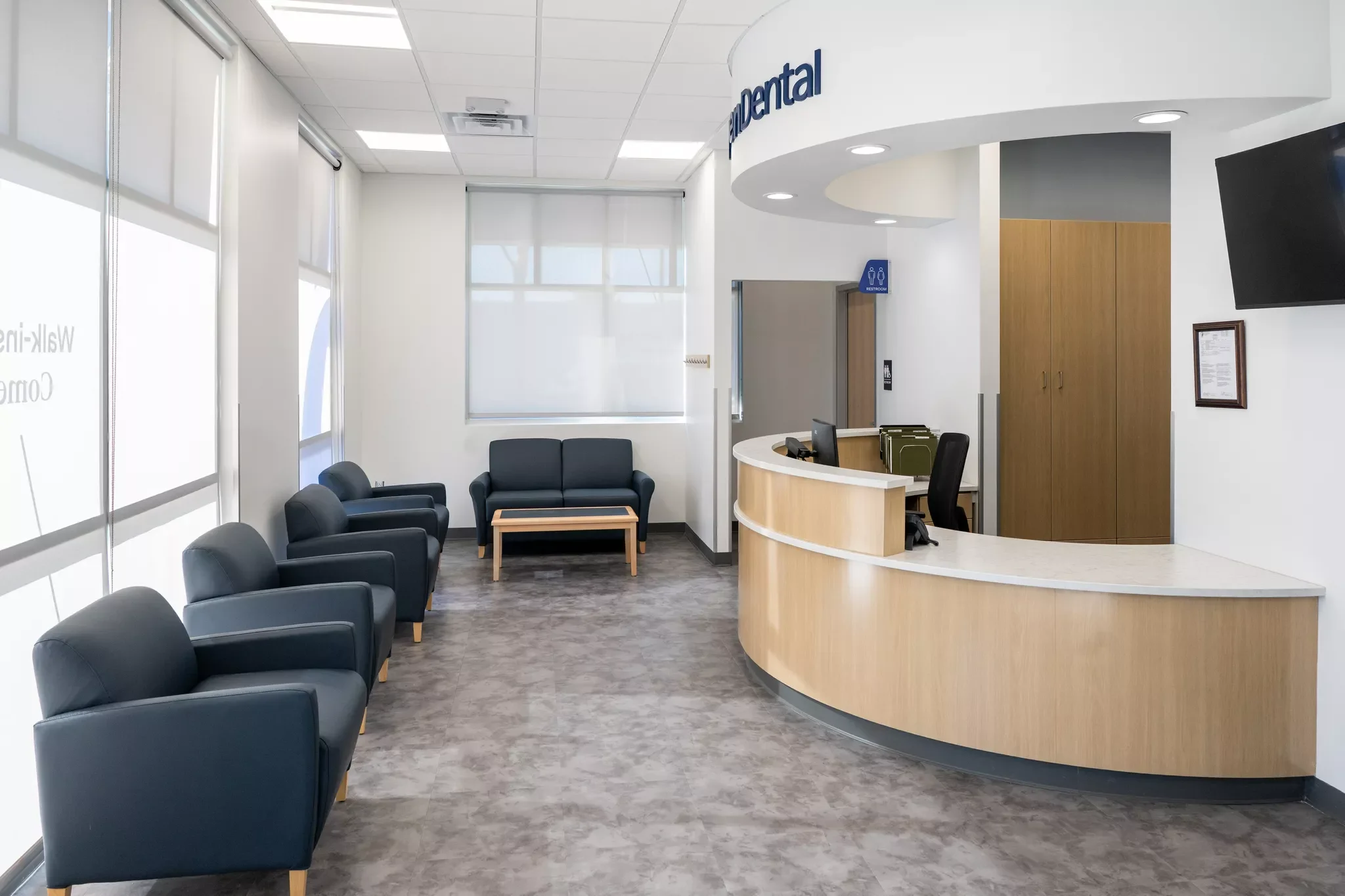 Empty dental office reception area with black chairs, a curved wooden reception desk, a television, and a sign indicating restrooms.