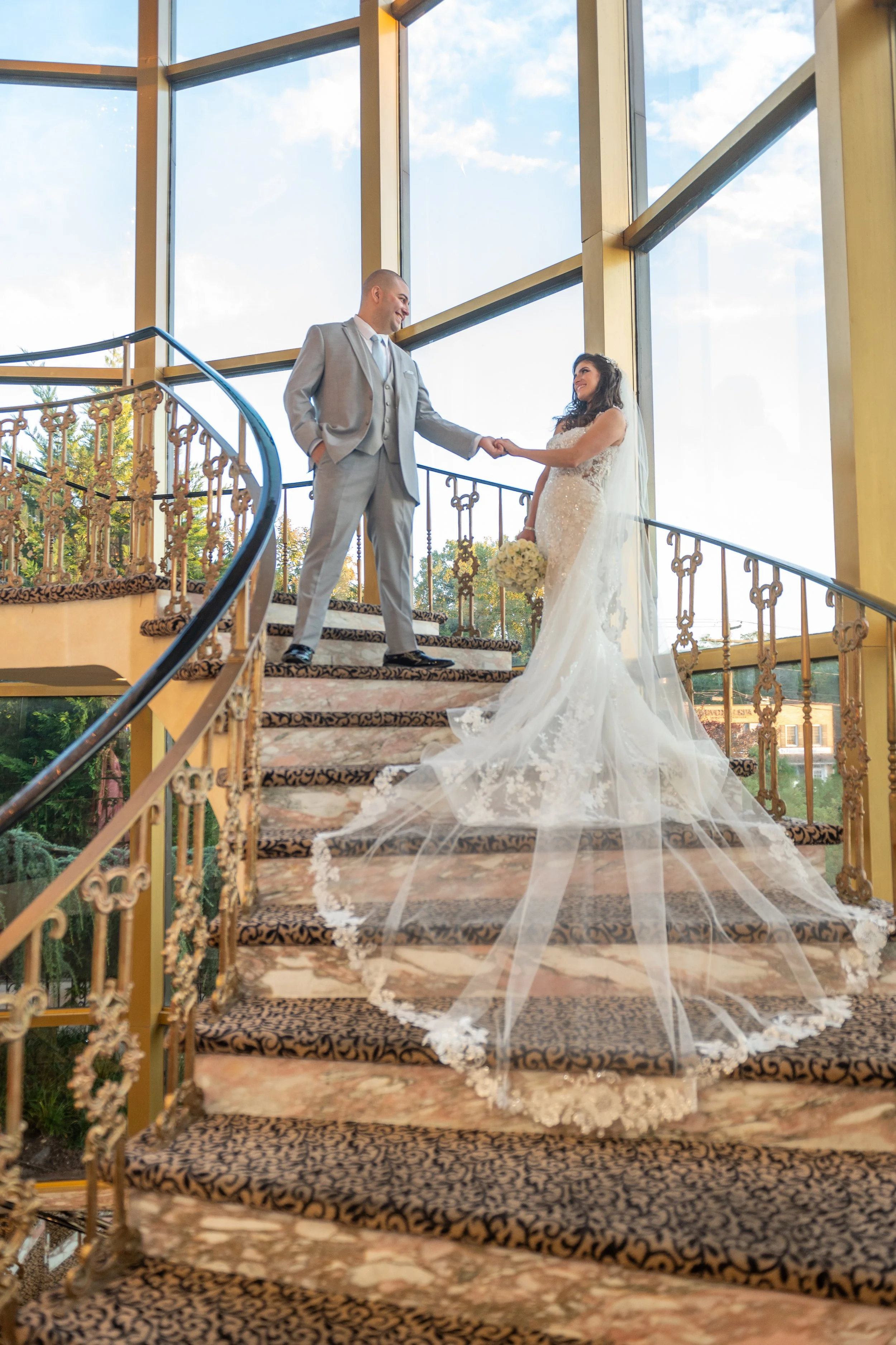A bride and groom on a staircase during their wedding ceremony, holding hands and looking at each other. The groom is dressed in a gray suit, and the bride is wearing a white wedding gown with a long veil and holding a bouquet.