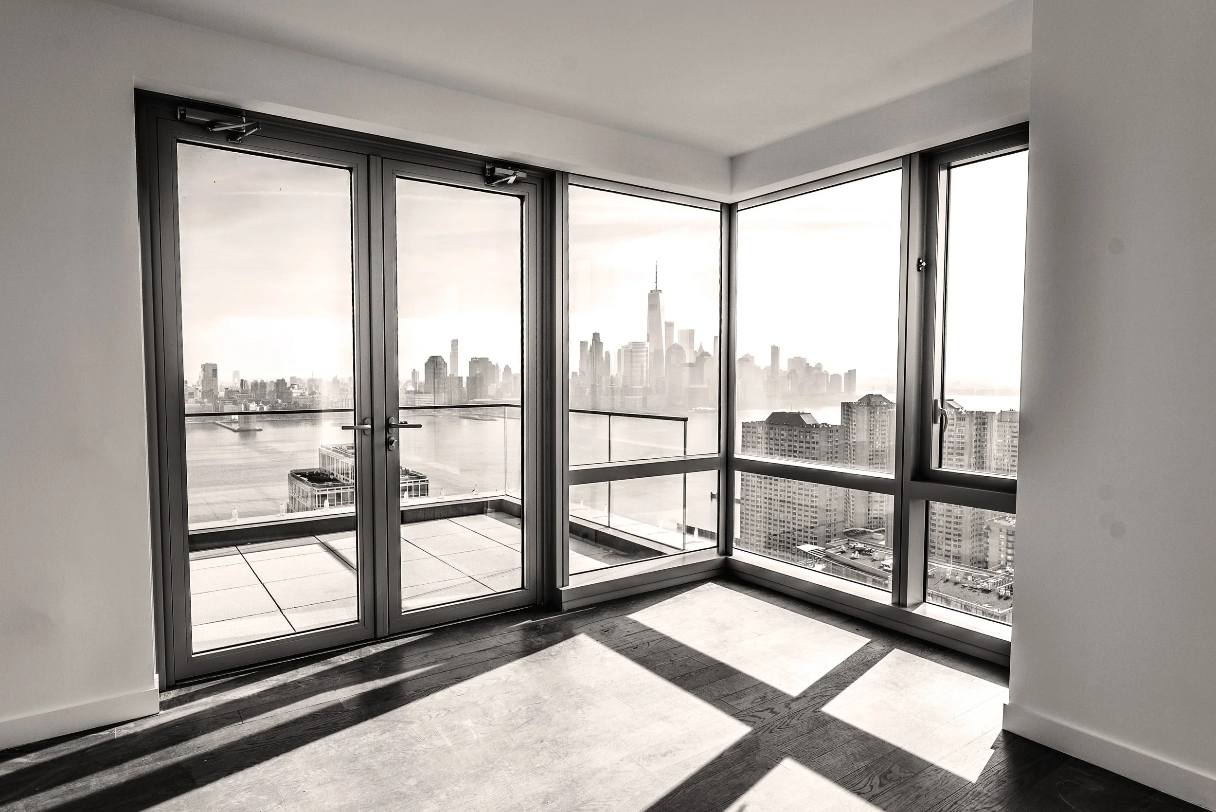 Empty room with large glass windows and door, overlooking a city skyline with skyscrapers and water, in black and white.