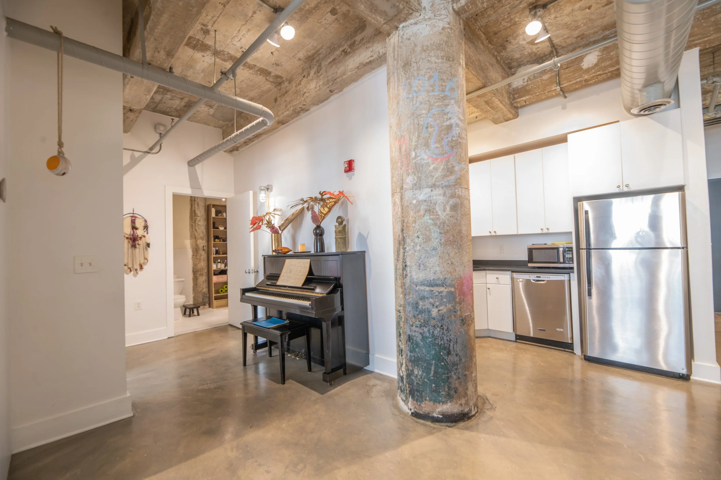 Interior view of a modern loft apartment with exposed concrete ceiling and a large support column. Features include a black upright piano with sheet music, decorative vases with dried flowers on top, a microwave, dishwasher, and stainless steel refrigerator in a white kitchen area.