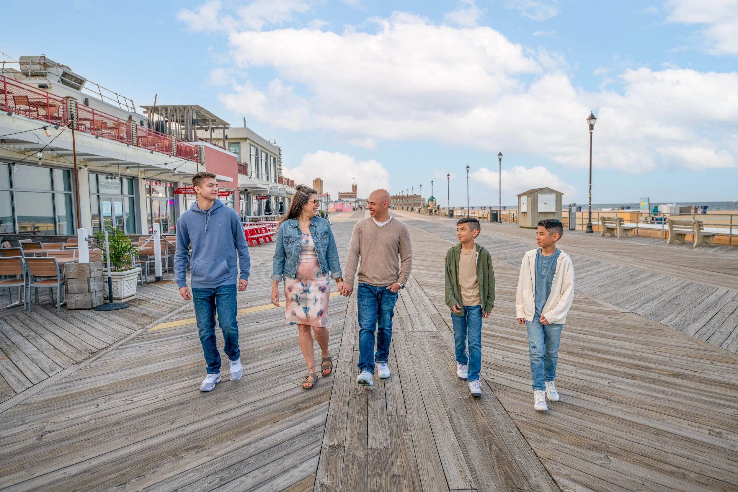 A family of six walking on a wooden pier near a beach, with shops and benches in the background under a partly cloudy sky.