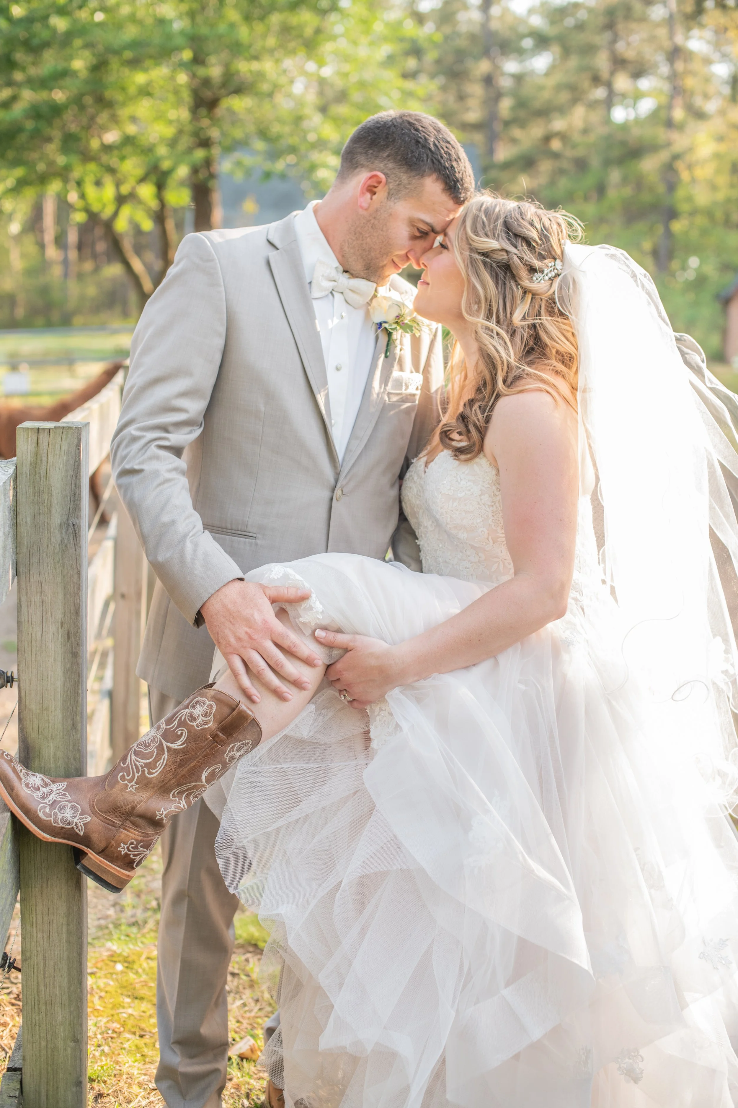A bride and groom in wedding attire sharing a tender moment outdoors, with the groom lifting the bride's wedding dress to reveal cowboy boots, in a forested setting.