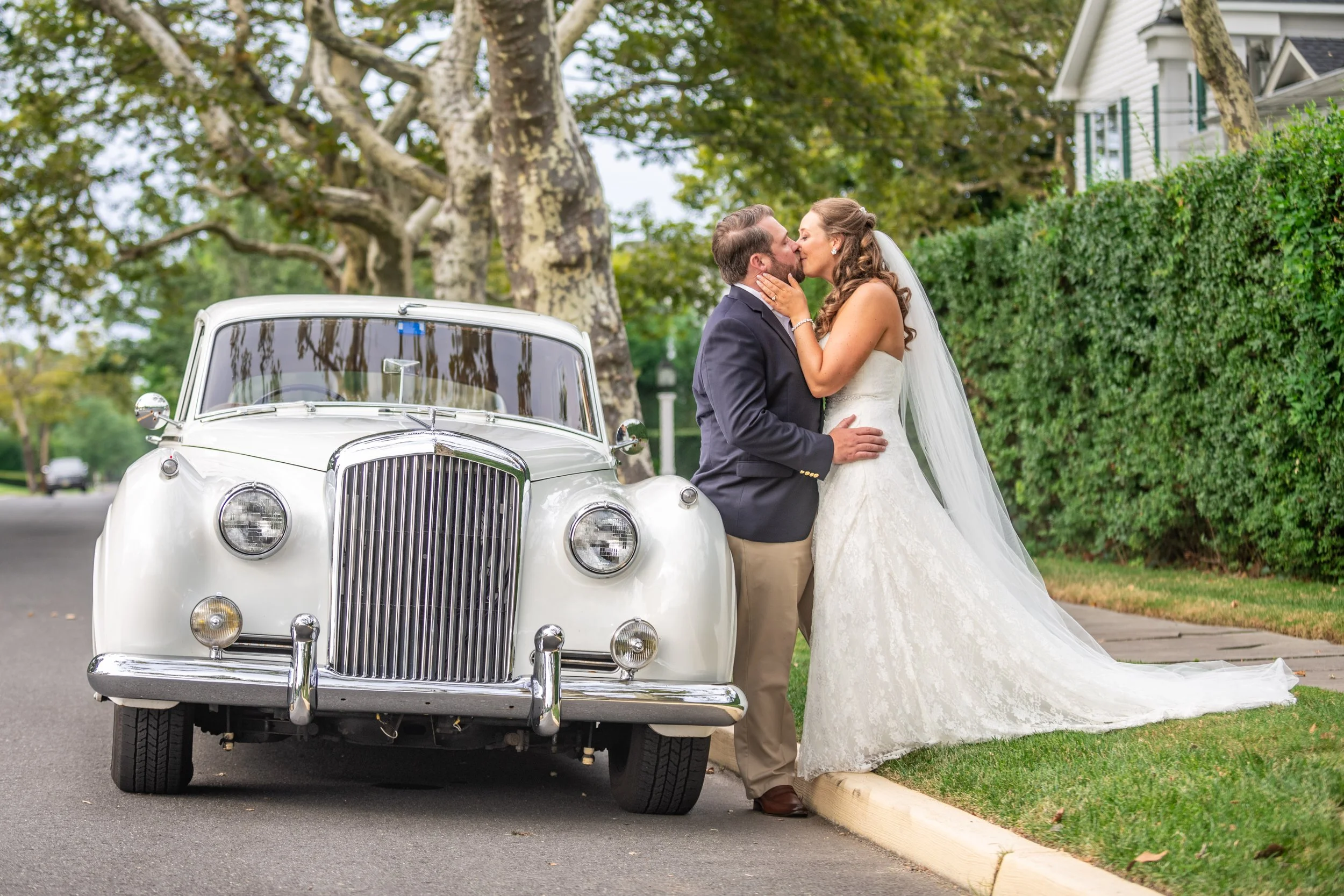 A bride and groom are sharing a kiss outdoors next to a vintage white car on a suburban street lined with trees and houses.