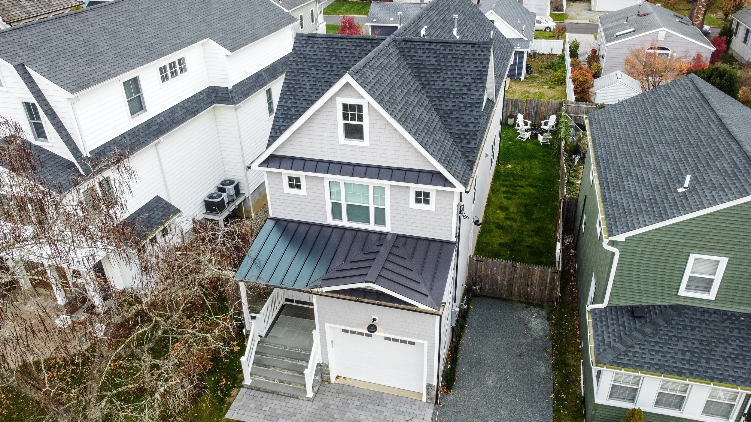 An aerial view of a modern house with a gray and black roof, white exterior, and a small front porch. The backyard features a lawn and a patio with outdoor chairs. The house is surrounded by neighboring houses with similar roofing and exterior colors.
