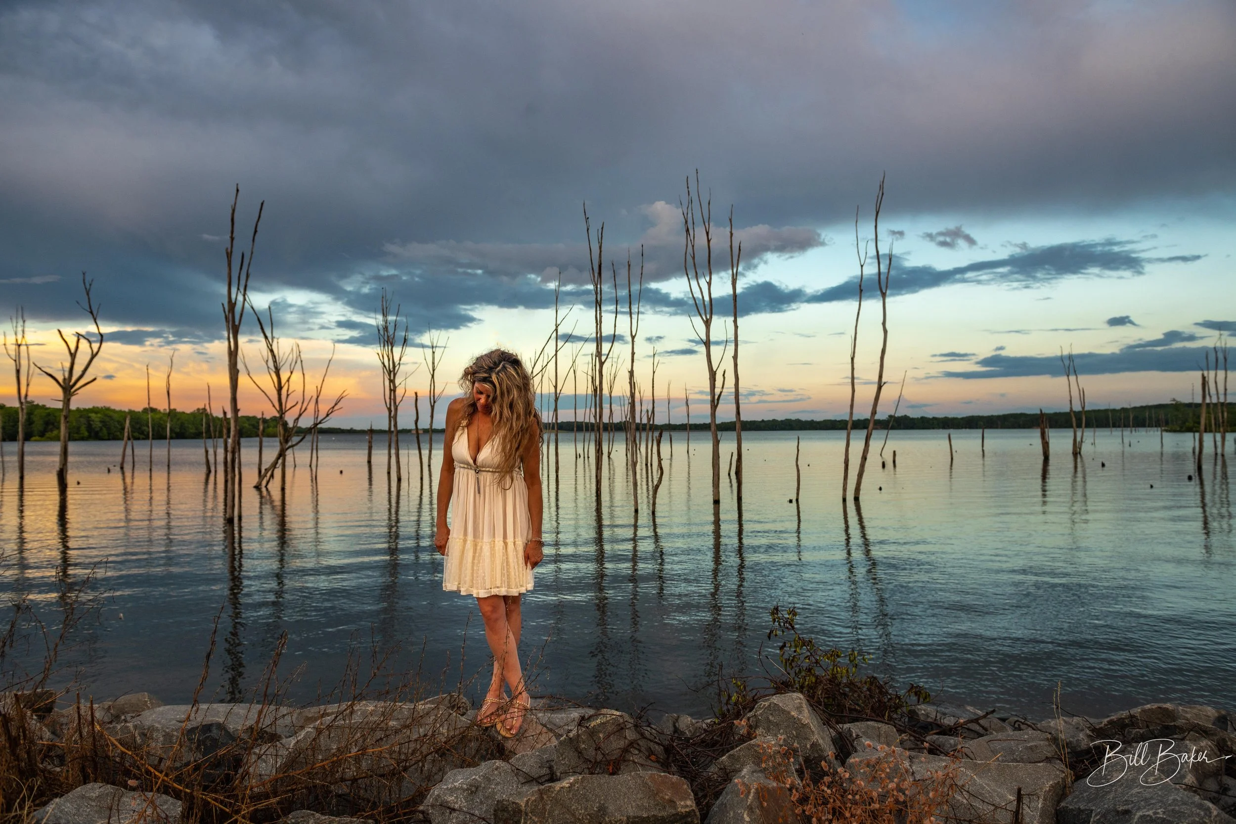 A woman in a white dress with wavy blonde hair standing on rocks by a lake at sunset, with dry trees sticking out of the water and a partly cloudy sky.