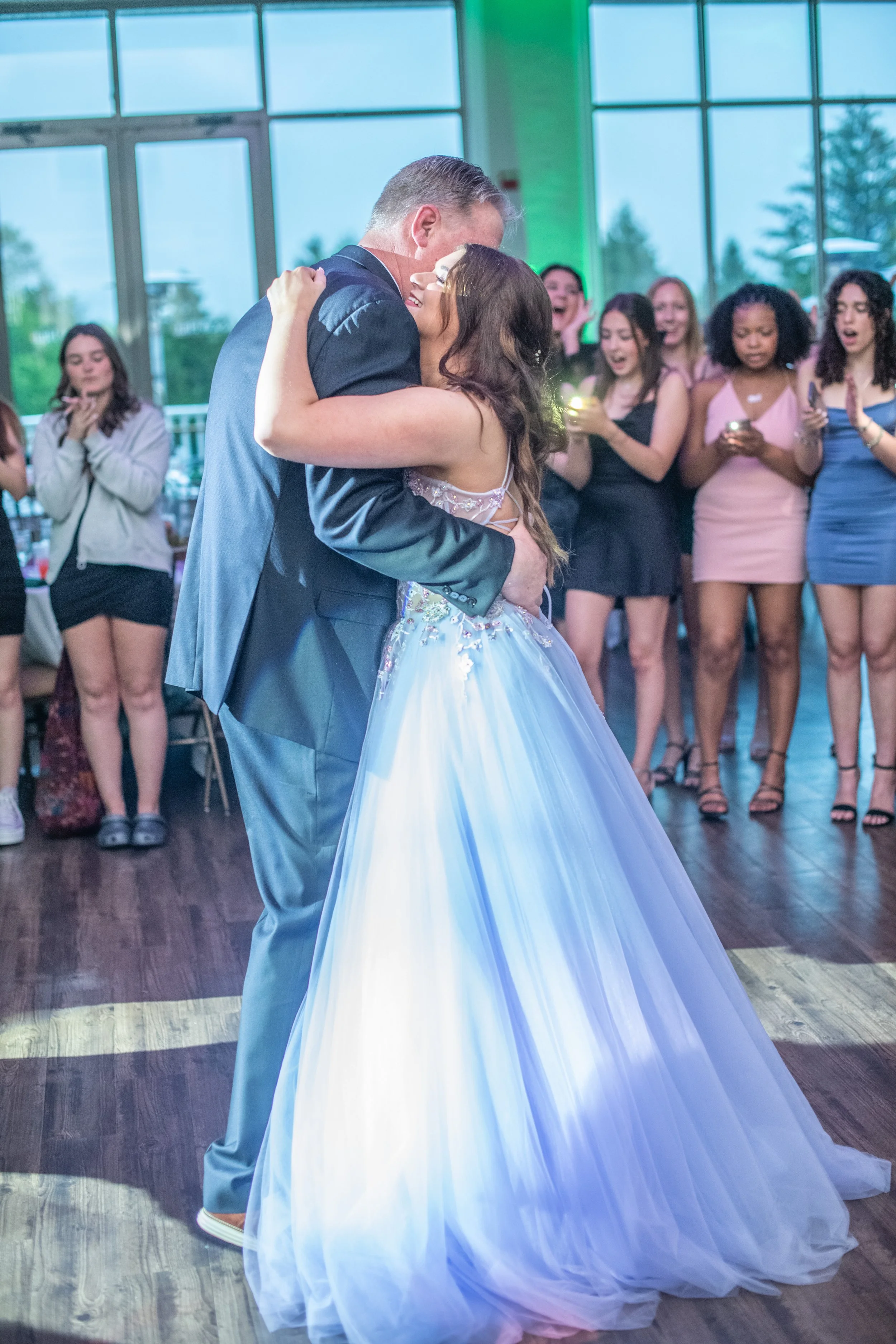 A couple dancing closely at their wedding reception with friends surrounding them, some taking photos, in a bright indoor setting.