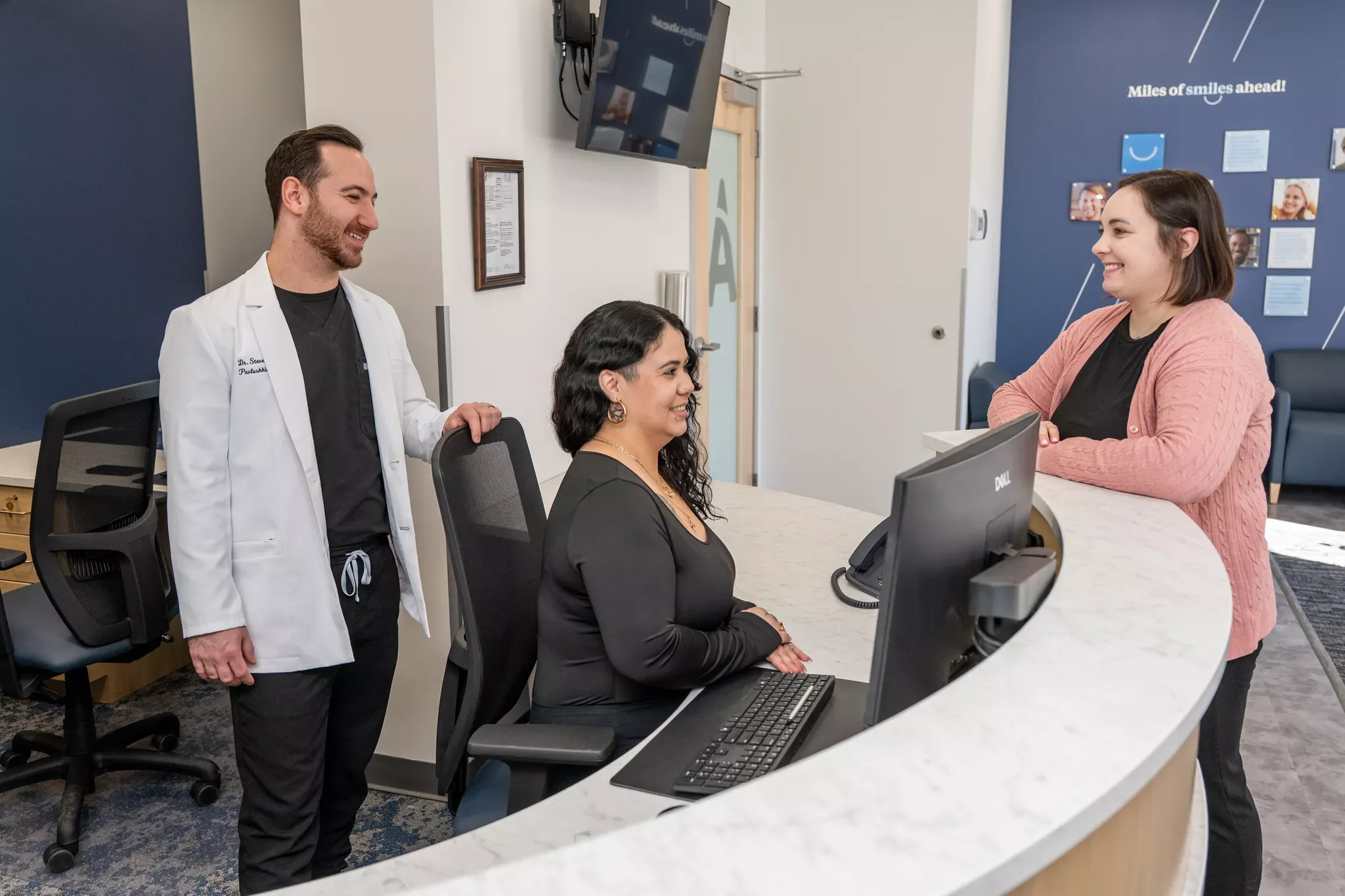 A woman at a reception desk smiling and talking to a woman standing in front of the desk, with a man doctor standing beside them in a medical office lobby.