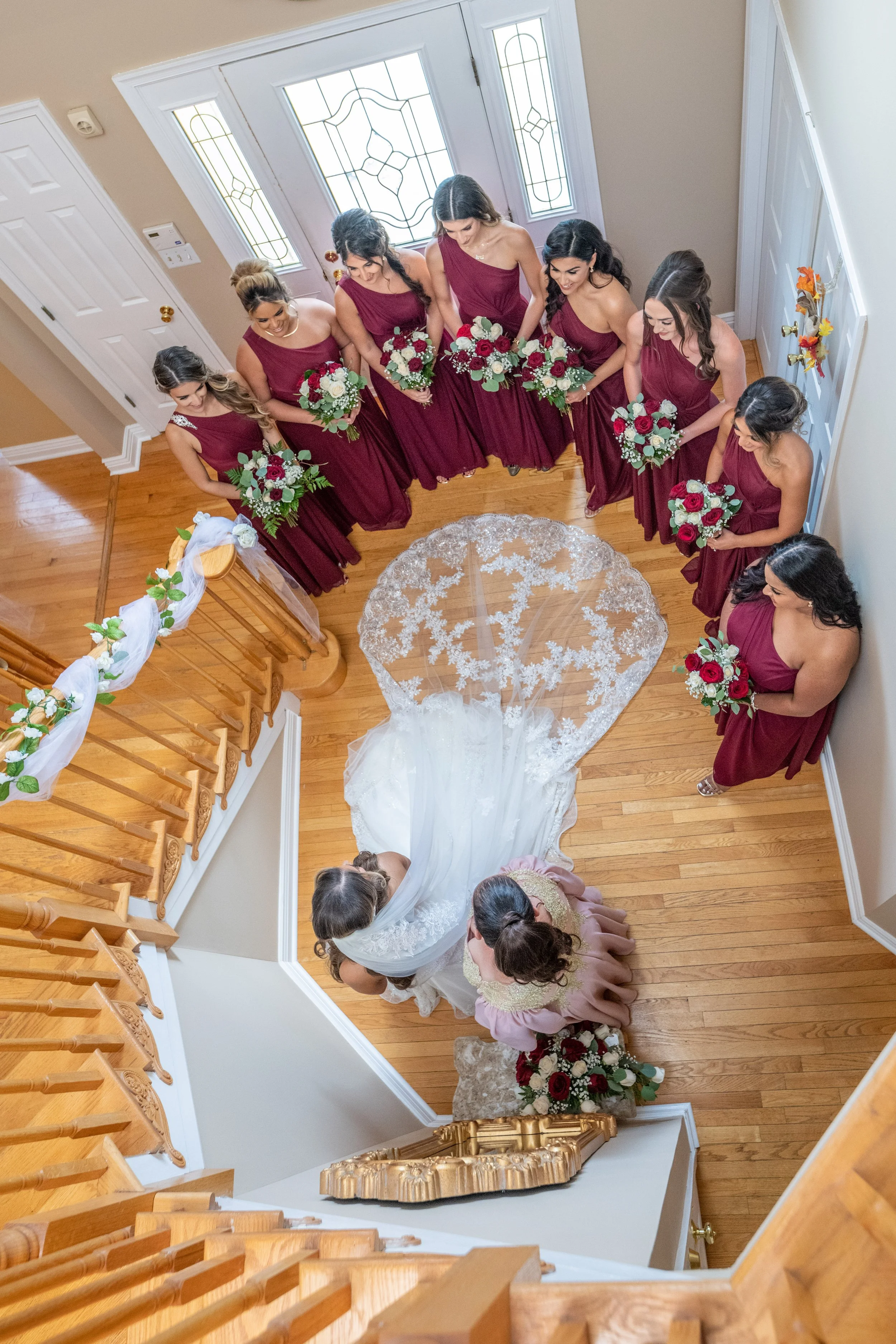 Bridal party standing around the top of a staircase during a wedding ceremony, with bridesmaids dressed in burgundy gowns holding bouquets, looking down at the bride and officiant below.