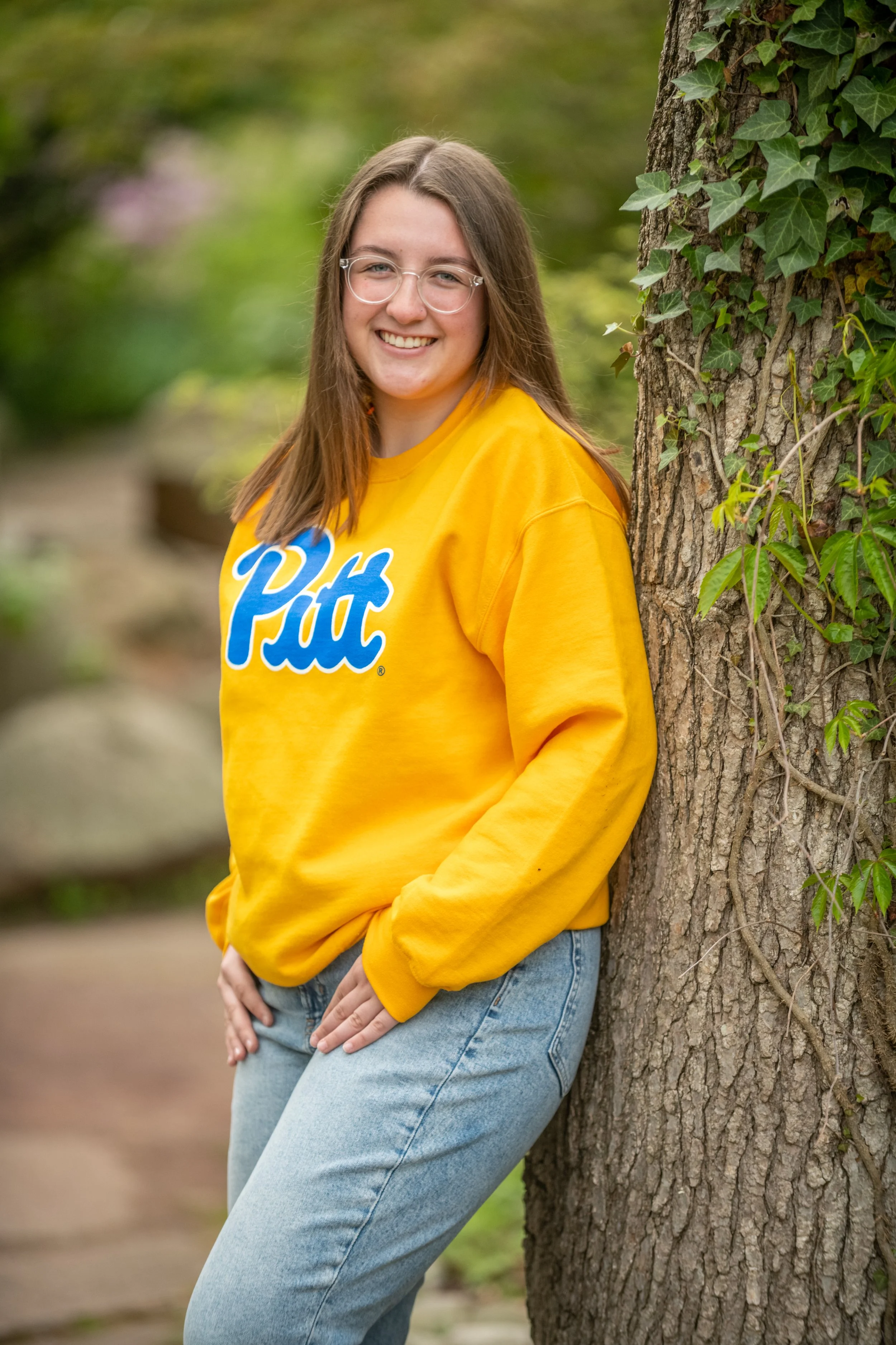 A young woman with glasses, wearing a yellow Pitt sweatshirt and light blue jeans, standing next to a tree outdoors with greenery in the background.