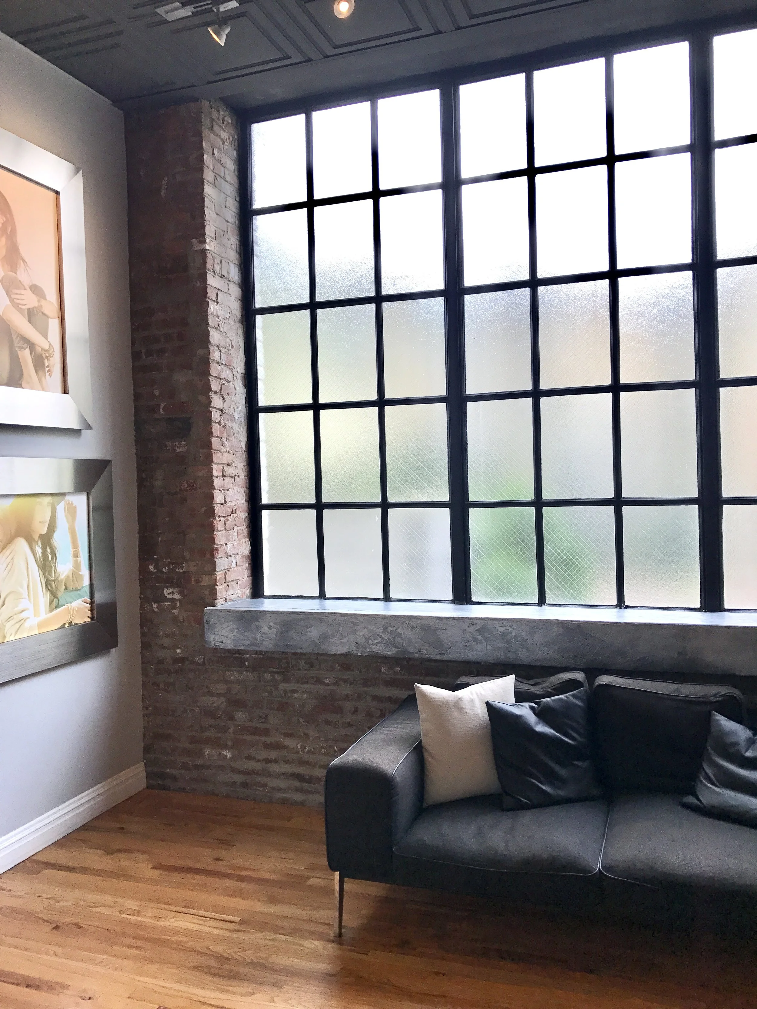 View of a modern living room with a large window composed of many small panes, a brick wall, a dark sofa with black and white pillows, and framed artwork on the wall.