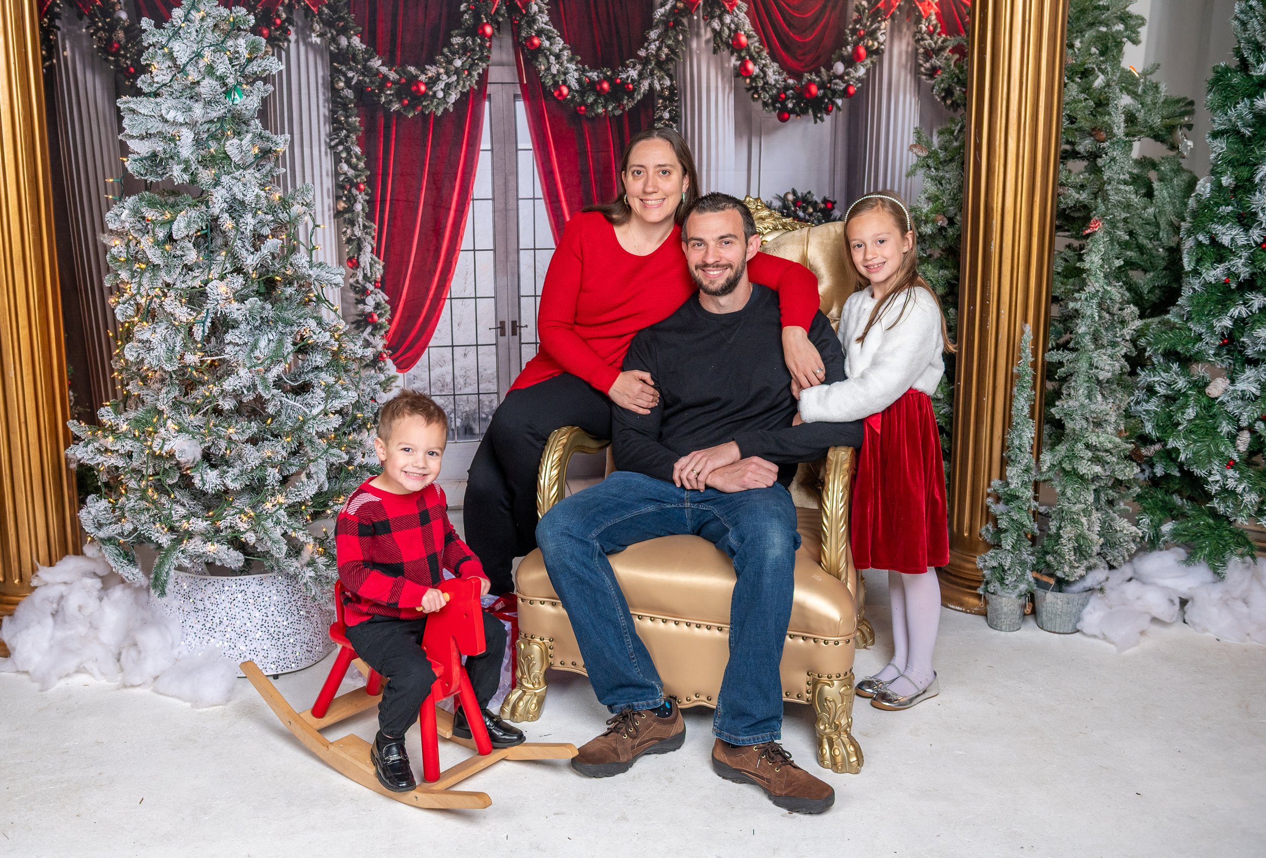 Family of five celebrating Christmas: a woman, a man seated on a golden throne with children around them, a boy on a red rocking horse, a girl in a red velvet dress, wintery scenery with snow-covered pine trees and Christmas decorations in the background.