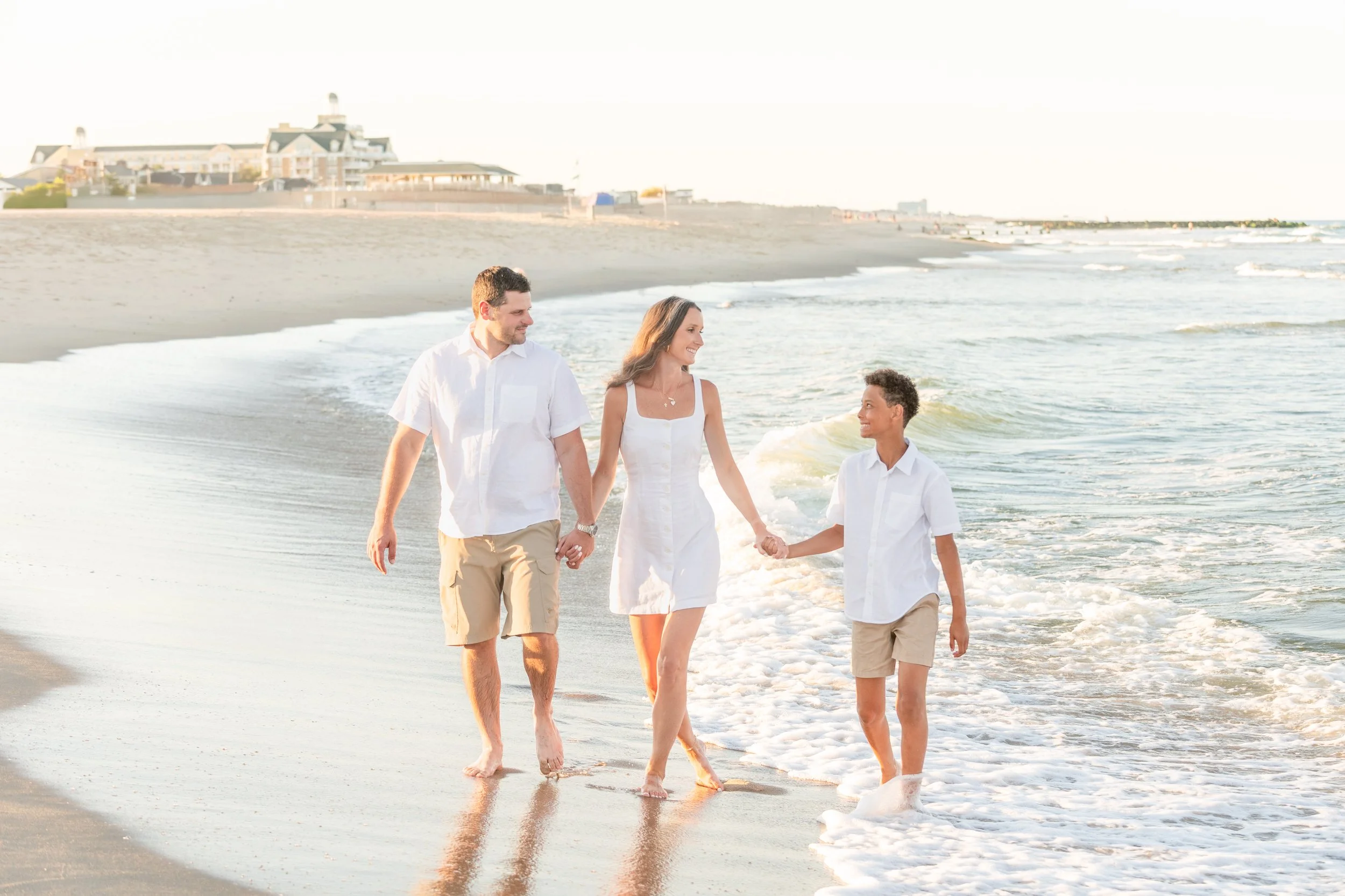 A family of three walking along the beach close to the shoreline, holding hands, with the ocean waves in front of them and houses visible in the background.