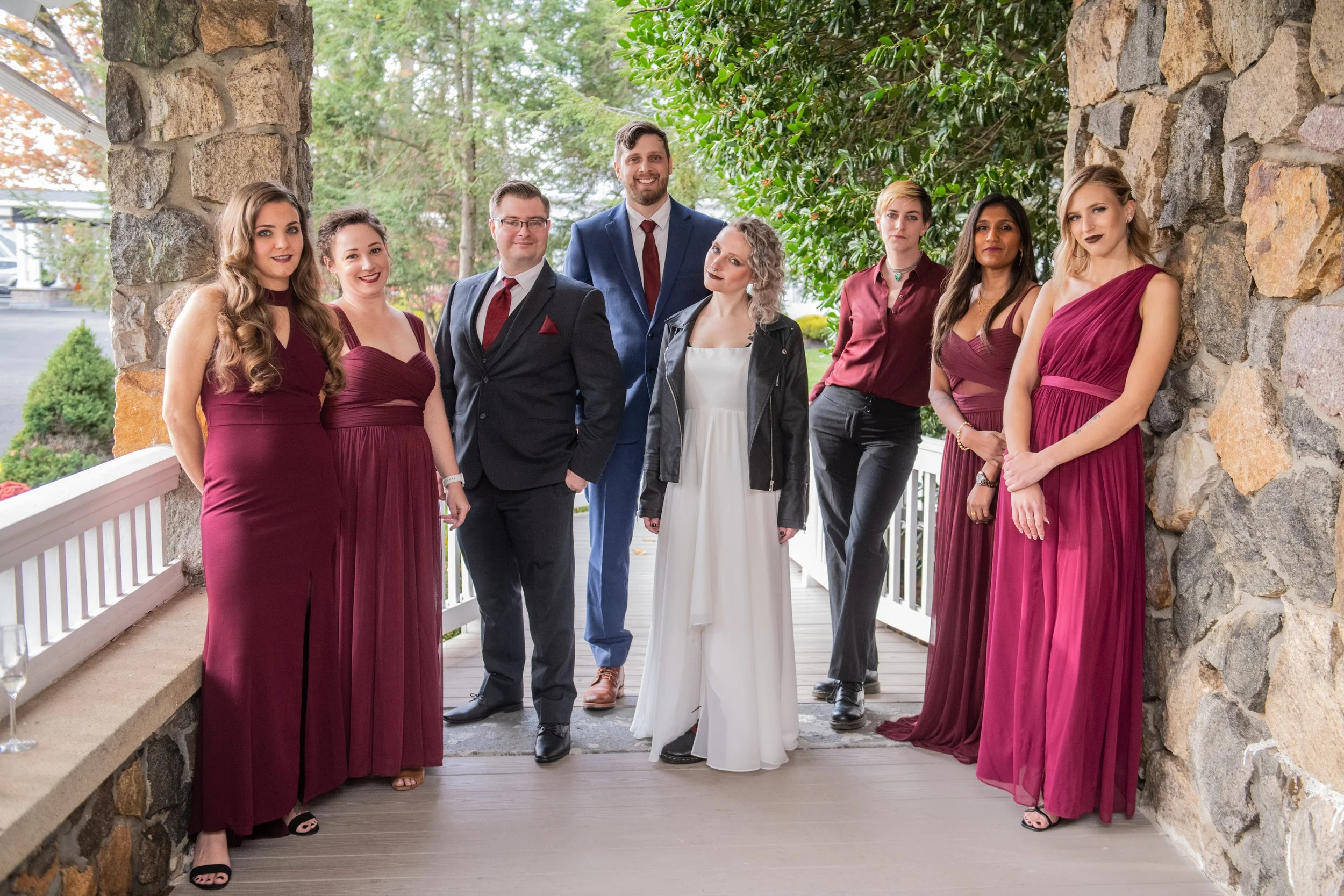 A group of nine people, consisting of men and women, standing on a porch with stone and wood elements. The women are wearing elegant burgundy dresses, and the men are dressed in suits. The background shows trees and a green outdoor setting.