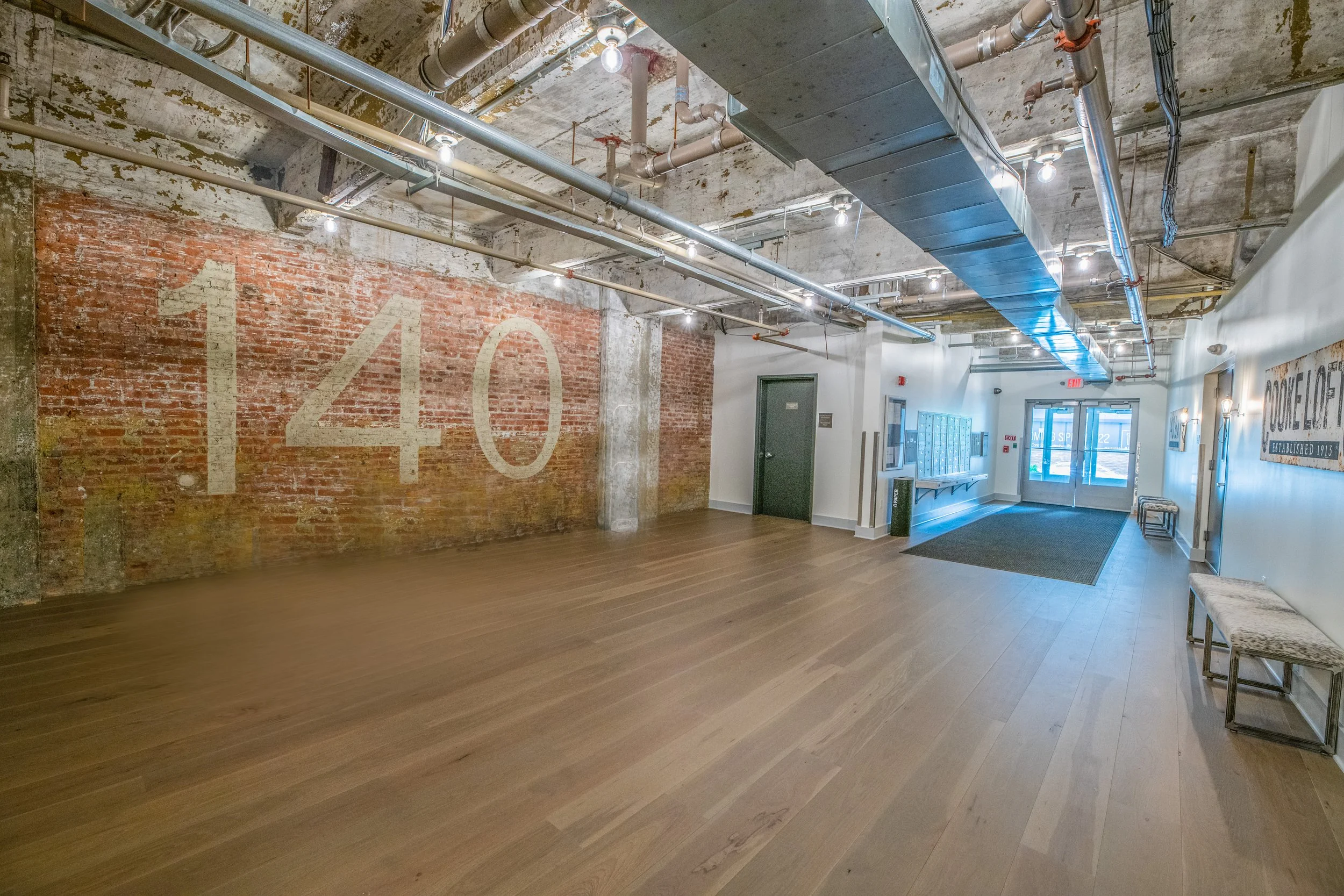 Empty interior space with a brick wall, painted numbers '140', exposed ceiling pipes, and hardwood flooring, possibly in a commercial or industrial building.