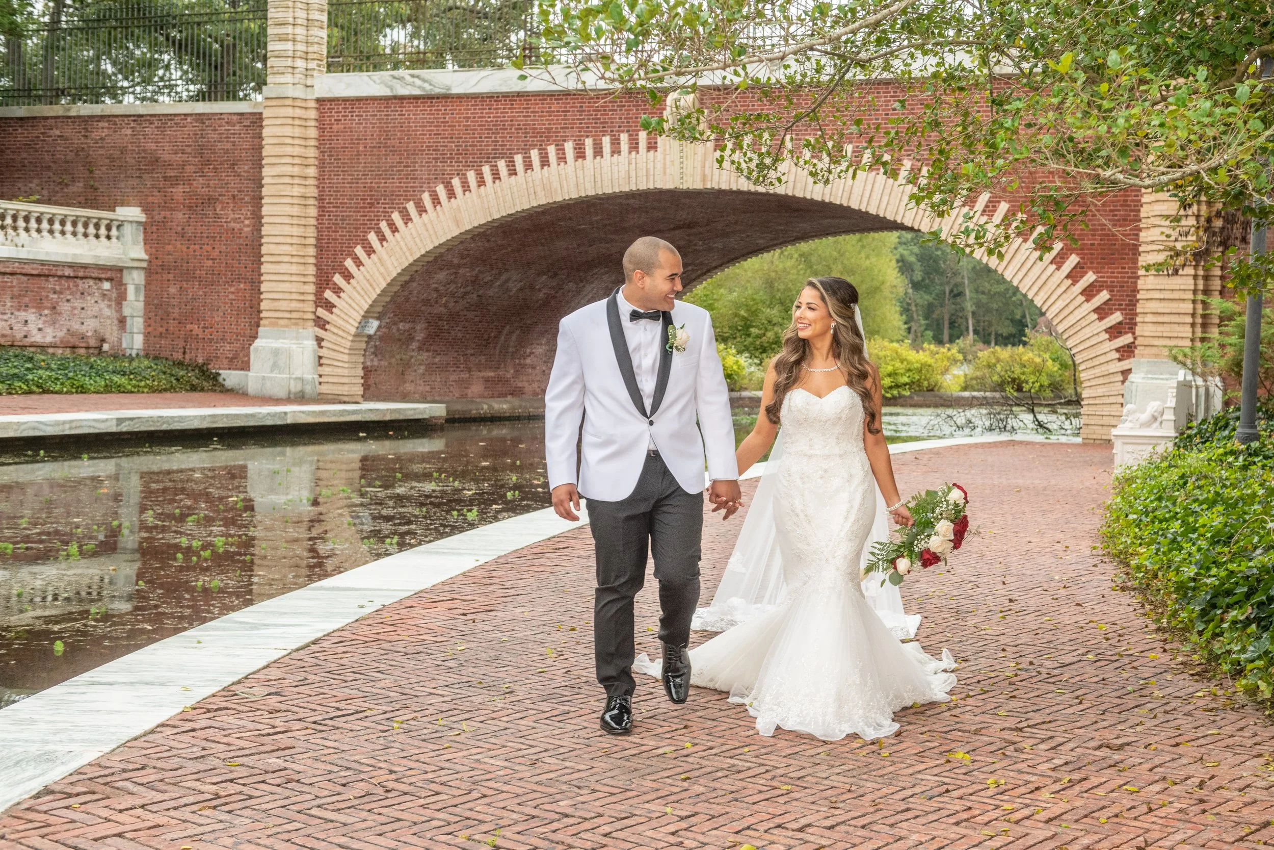 A newlywed couple walking hand in hand on a brick pathway near a pond, with an arched brick bridge and green trees in the background.