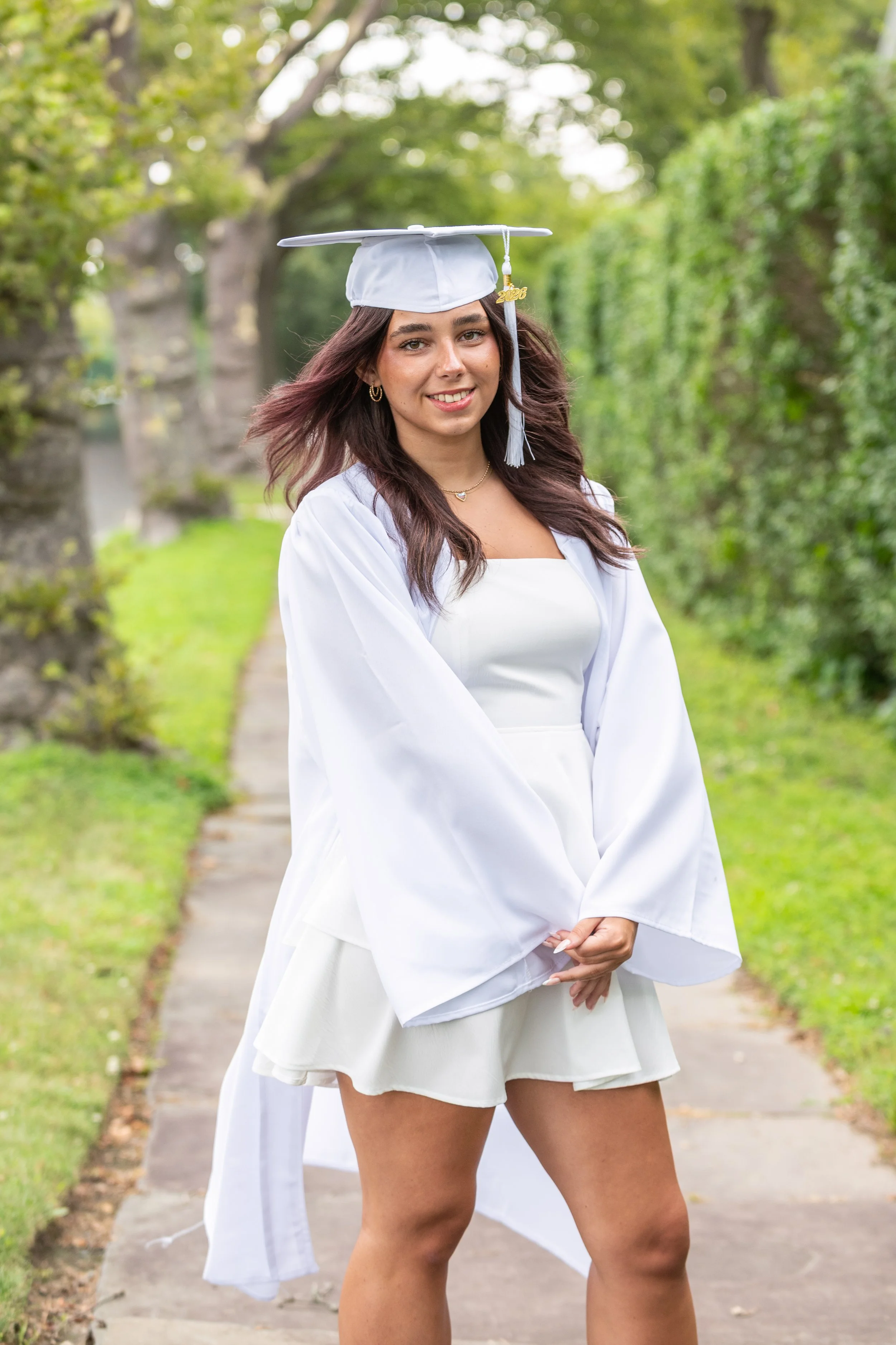 A young woman in a white graduation gown and cap standing on a sidewalk in a green park.