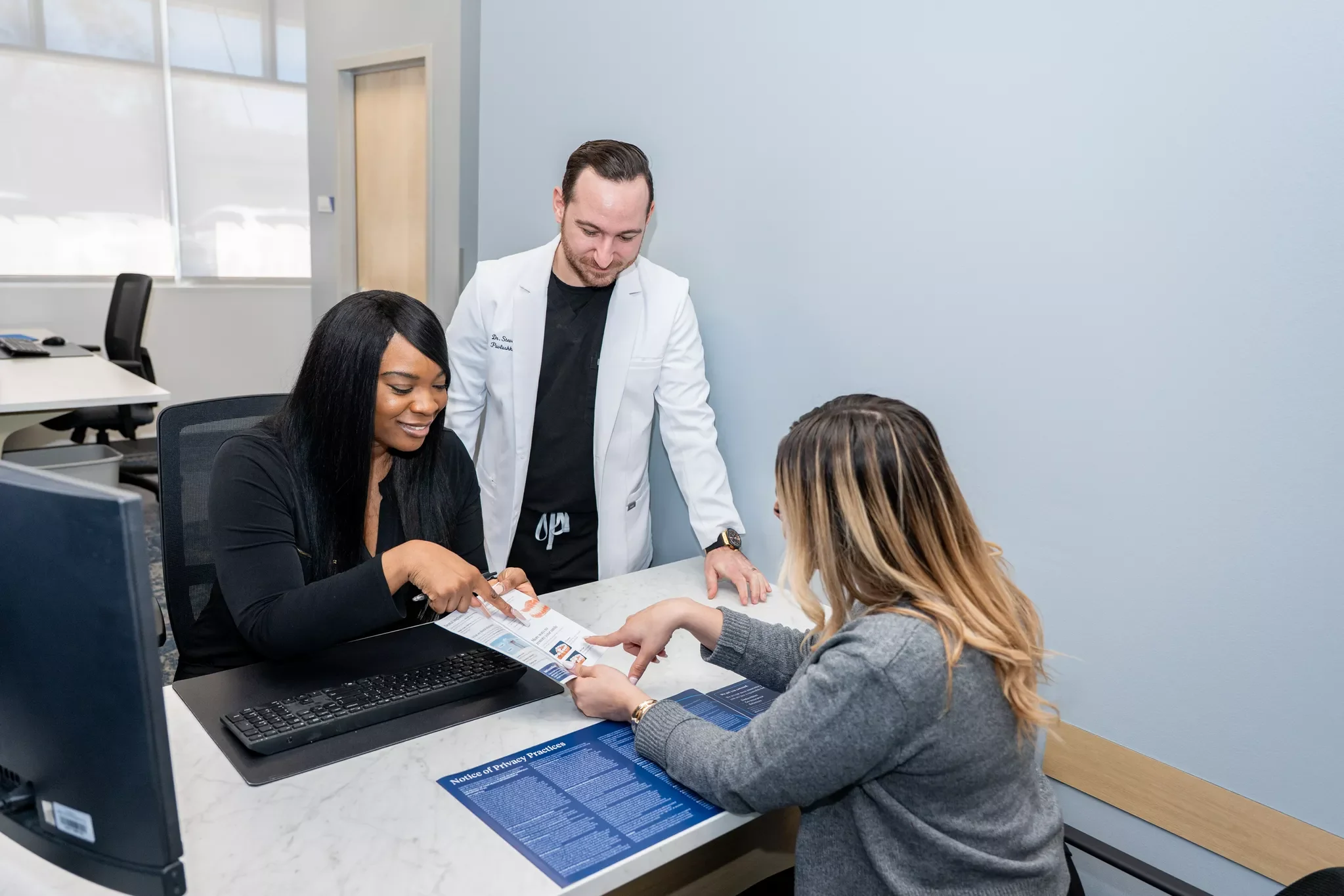 A woman and a man, possibly doctors, are sitting at a desk with a patient. The woman is pointing at a brochure, the patient is holding a sheet of paper, and the man is standing and looking at the documents, in an office setting.