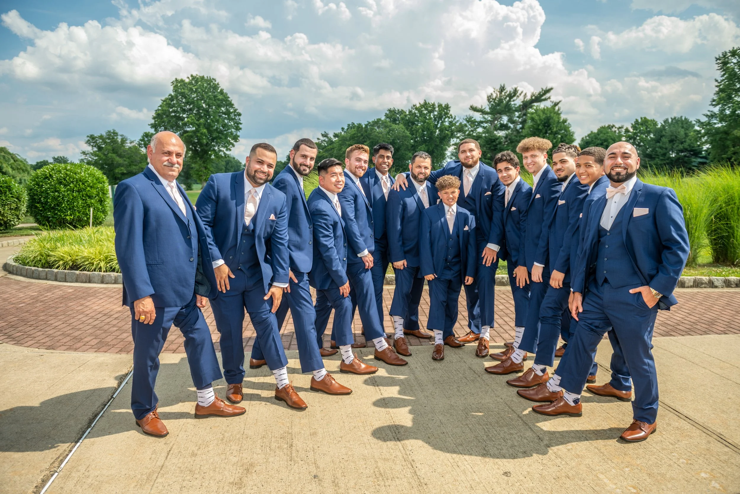 Group of men in blue suits and brown shoes, standing outdoors on a brick path, with greenery and trees in the background, smiling at the camera.