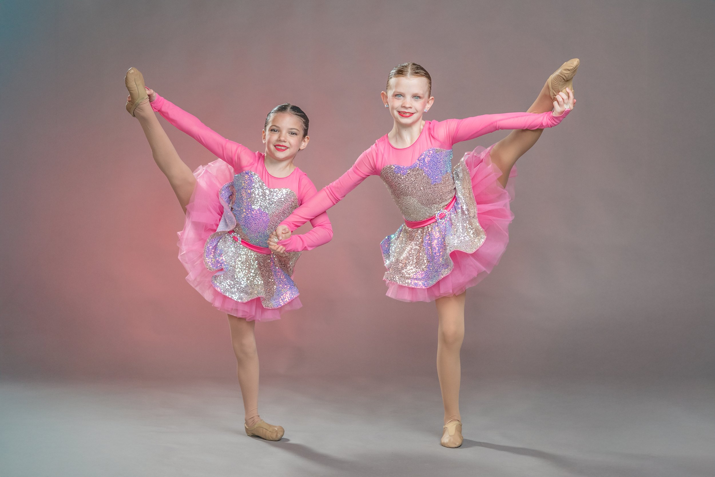 Two young girls in pink dance costumes performing ballet poses with one leg high in the air, smiling against a neutral background.