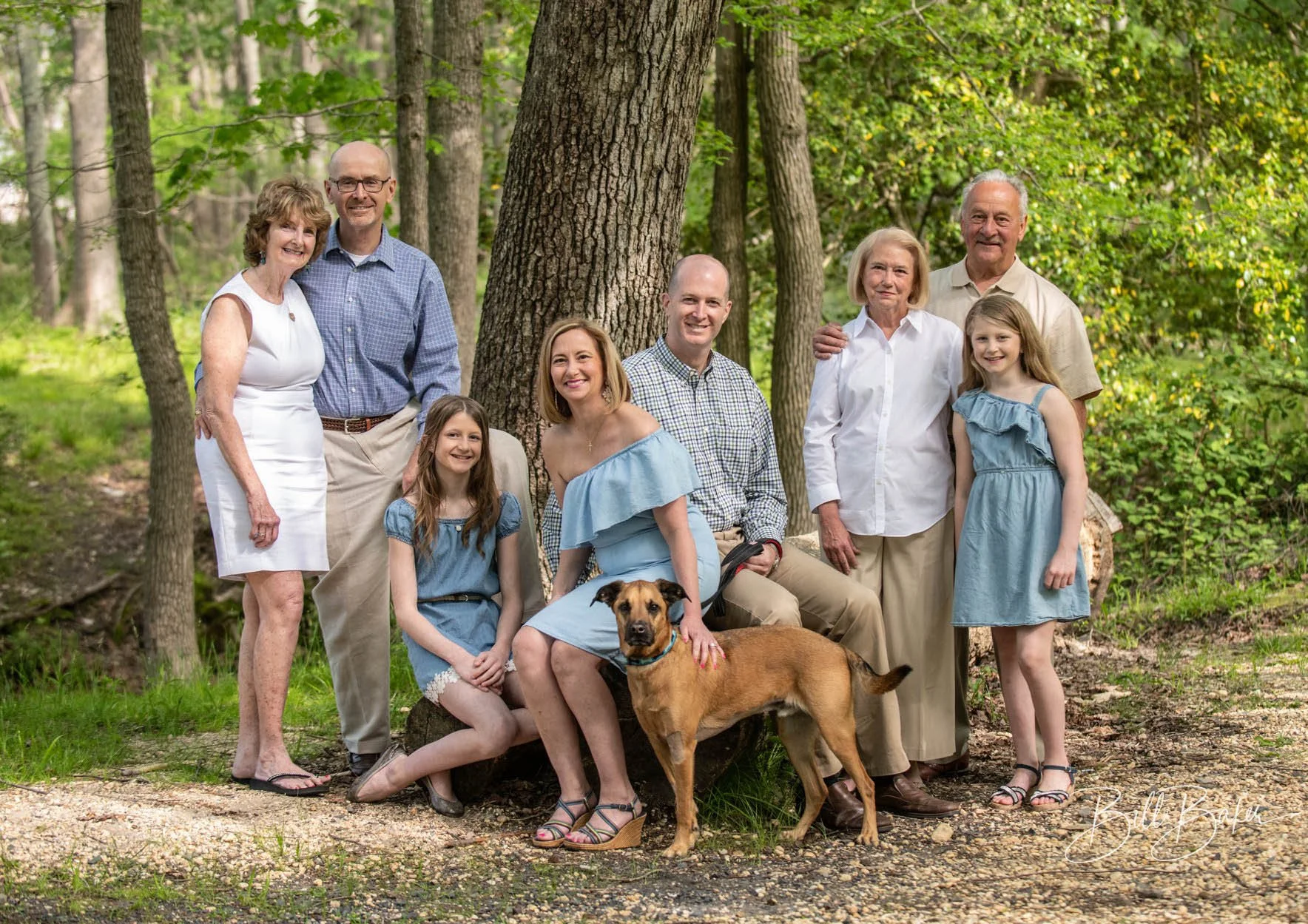 Family group photo outdoors in a wooded park, featuring eight people and a dog. The group includes three elderly women and men, two young girls, and two middle-aged women and men, all casually dressed and smiling.