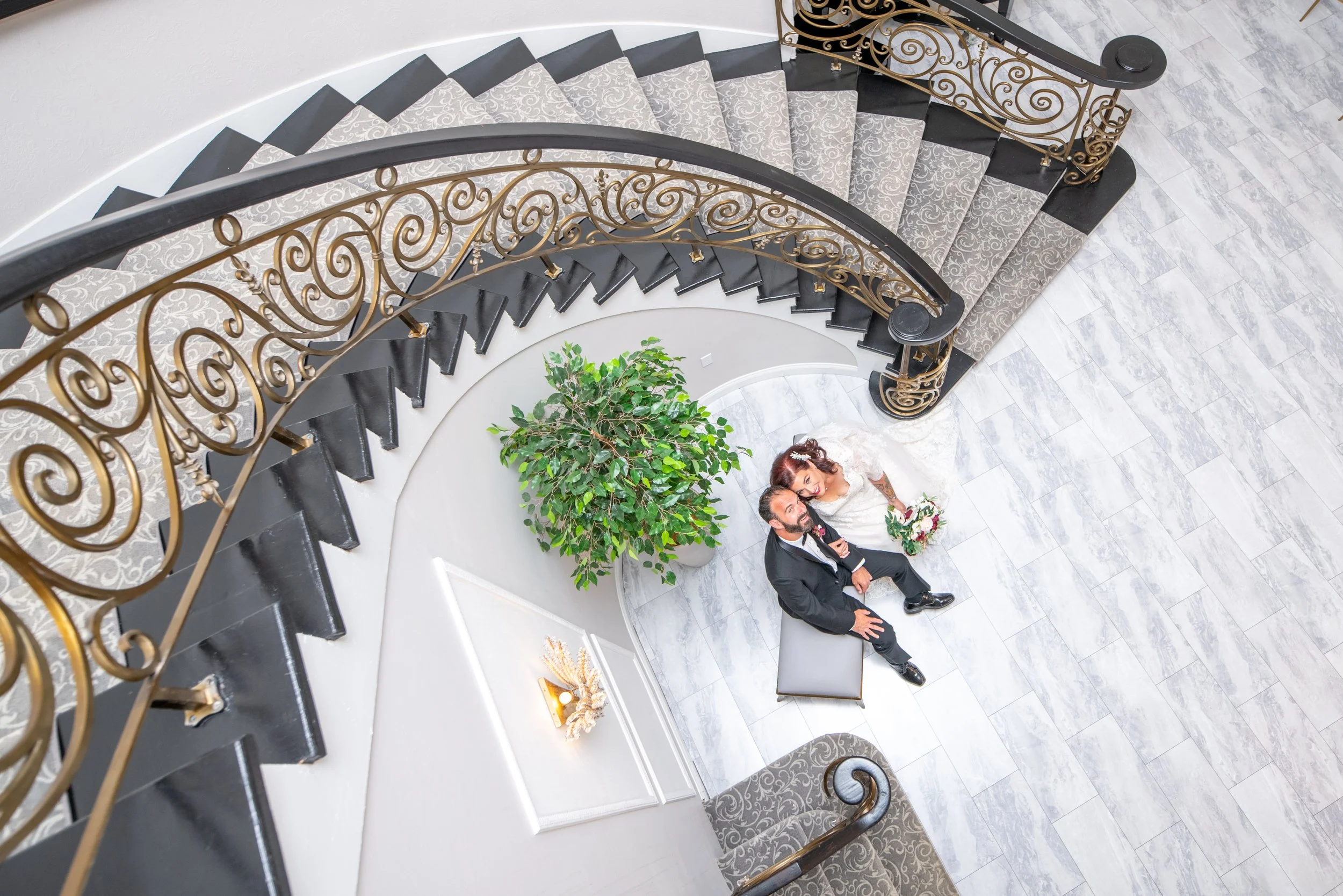 Overhead view of a bride and groom sitting on a bench beneath a decorative staircase with ornate gold railing, a potted green plant, and a light-colored tiled floor.