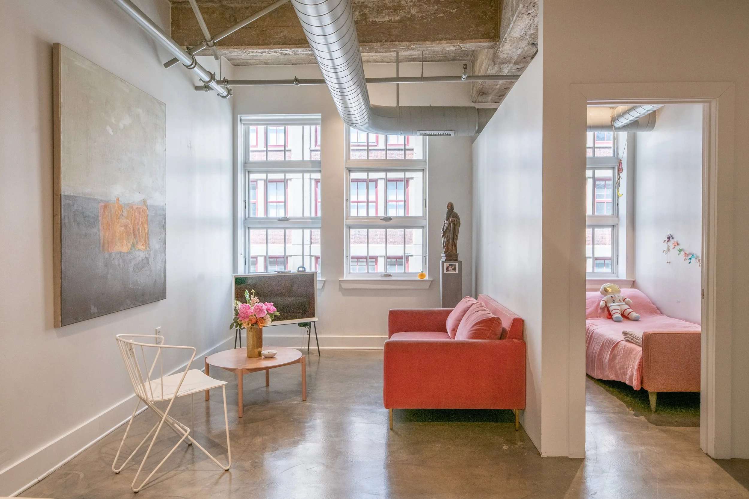 A living room with large windows, a pink sofa, a small table with a flower arrangement, and a white chair. There's a doorway leading to a bedroom with a pink bed, a doll, and window with natural light.