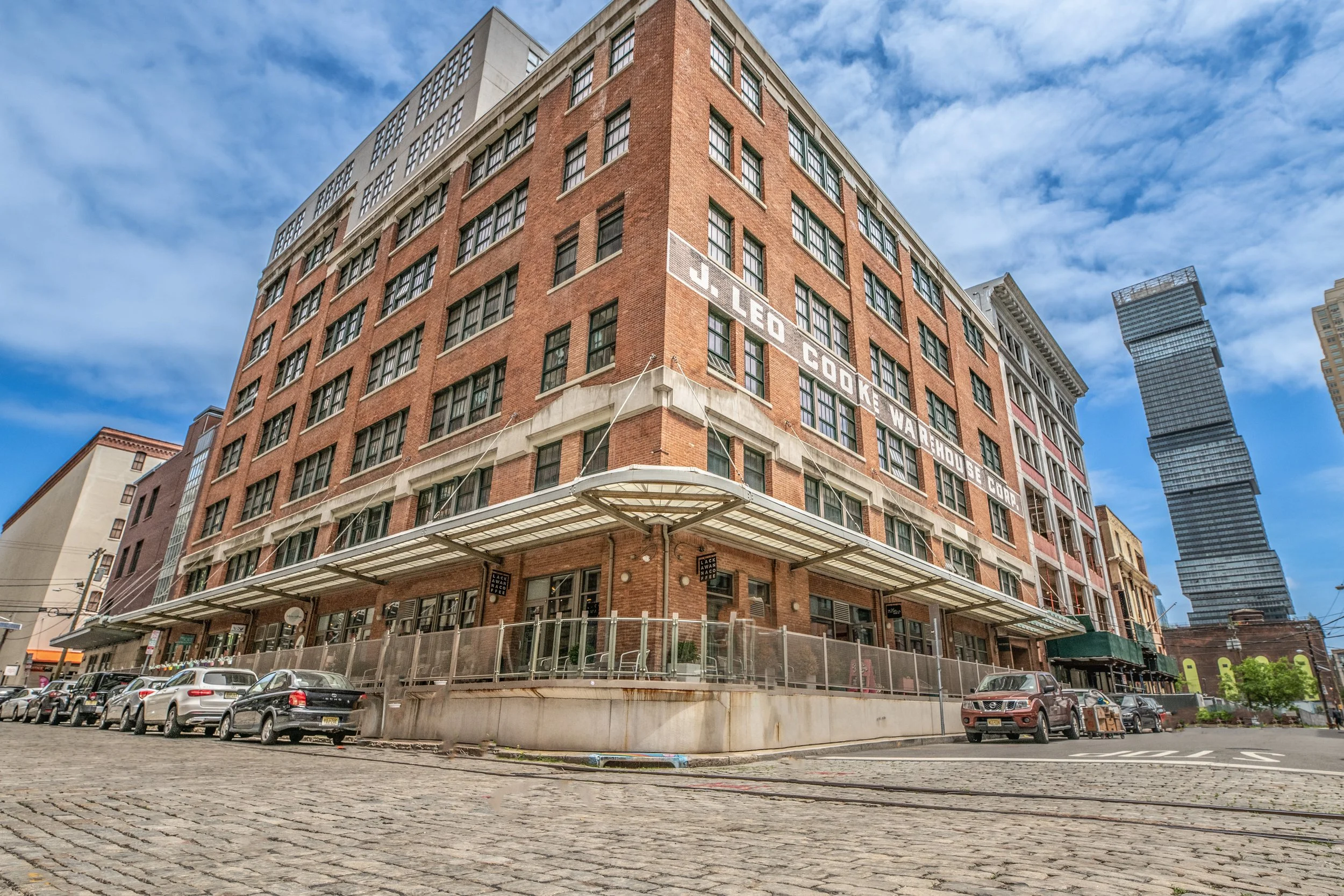 A large brick building with multiple floors and large windows, with a sign that reads, 'J. Leo Cookie Warehouse Corp.' on the side, situated on a cobblestone street with parked cars. The sky is partly cloudy, and a modern skyscraper is visible in the background.