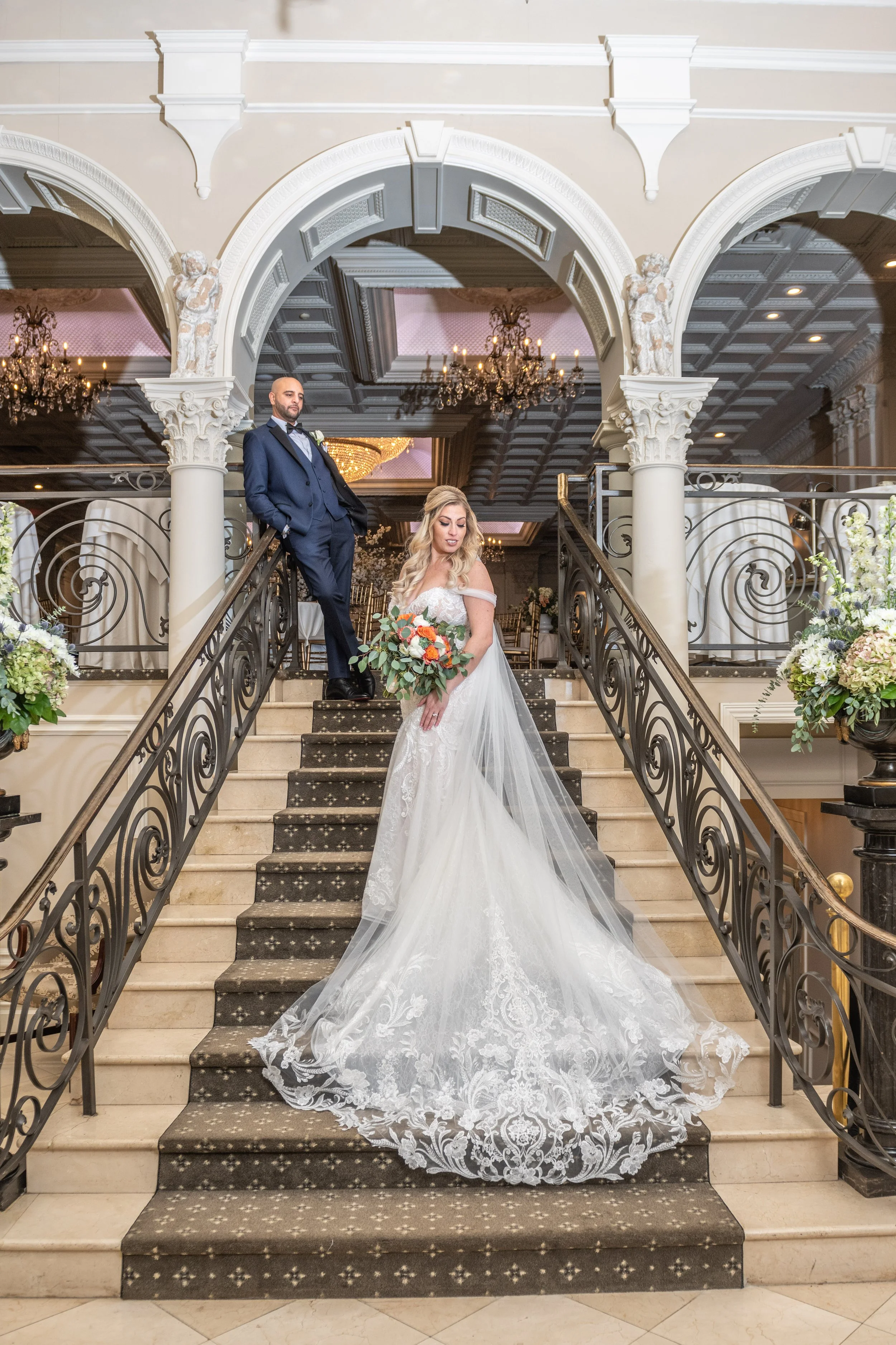 Bride in a white lace wedding gown holding a bouquet, standing on staircase with ornate wrought iron railings, with groom in a navy suit and bow tie standing behind her in an elegant indoor wedding venue with chandeliers and floral arrangements.