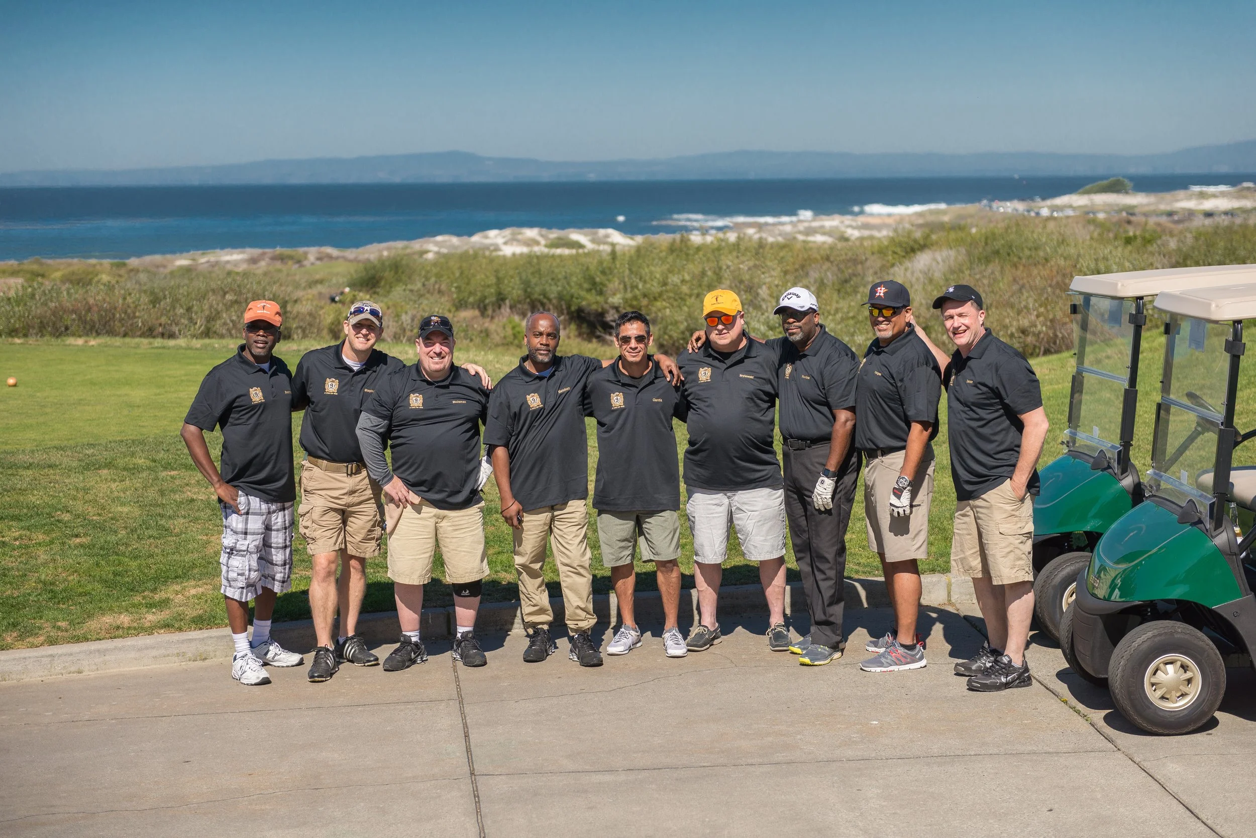 Group of nine men standing on a golf course near the beach, with ocean and hills in the background, all wearing black polo shirts, some with caps or sunglasses.