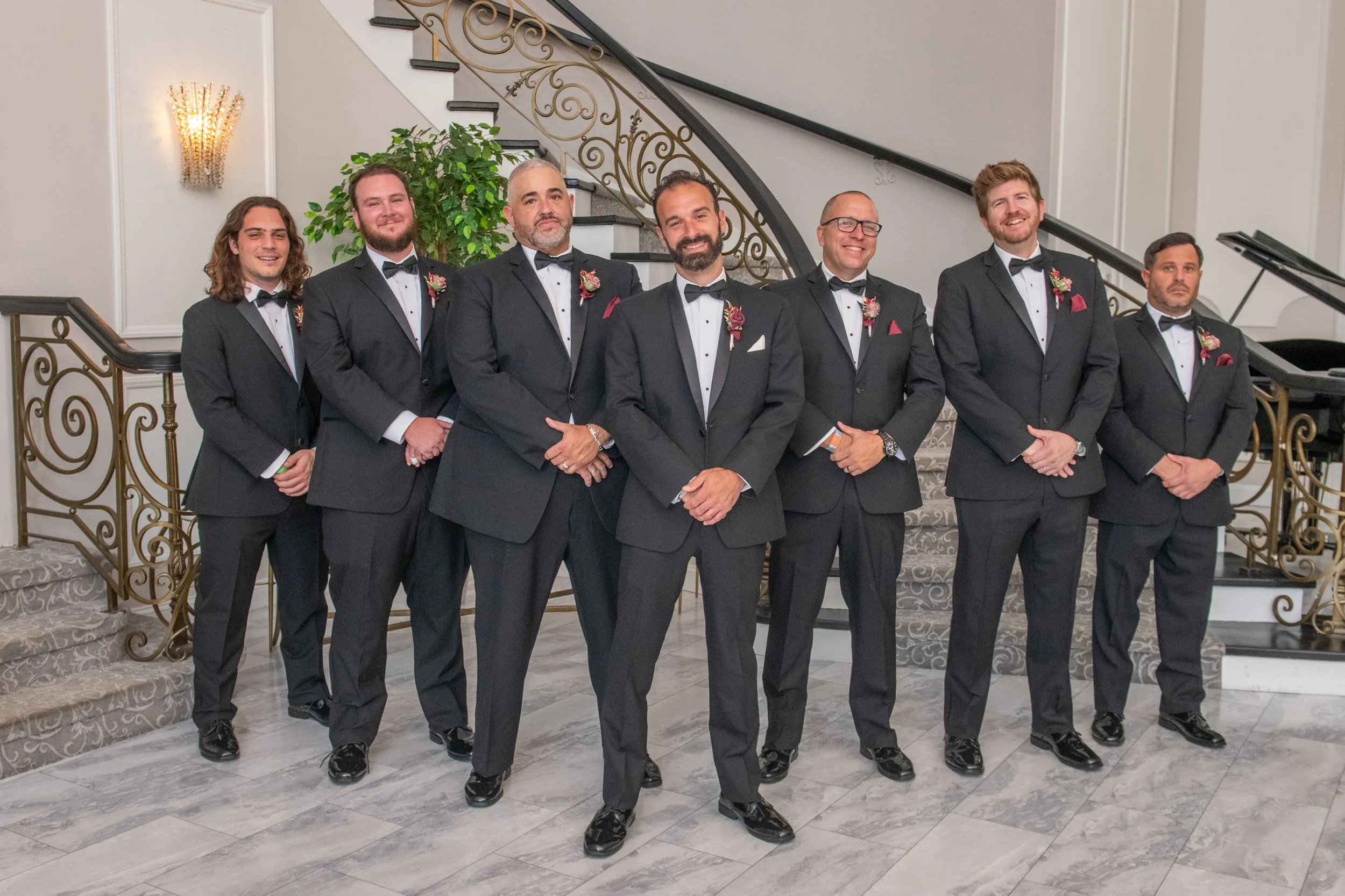 Seven men dressed in black tuxedos with bow ties, standing in a line indoors in front of a staircase with ornate gold railings, smiling for the photo.